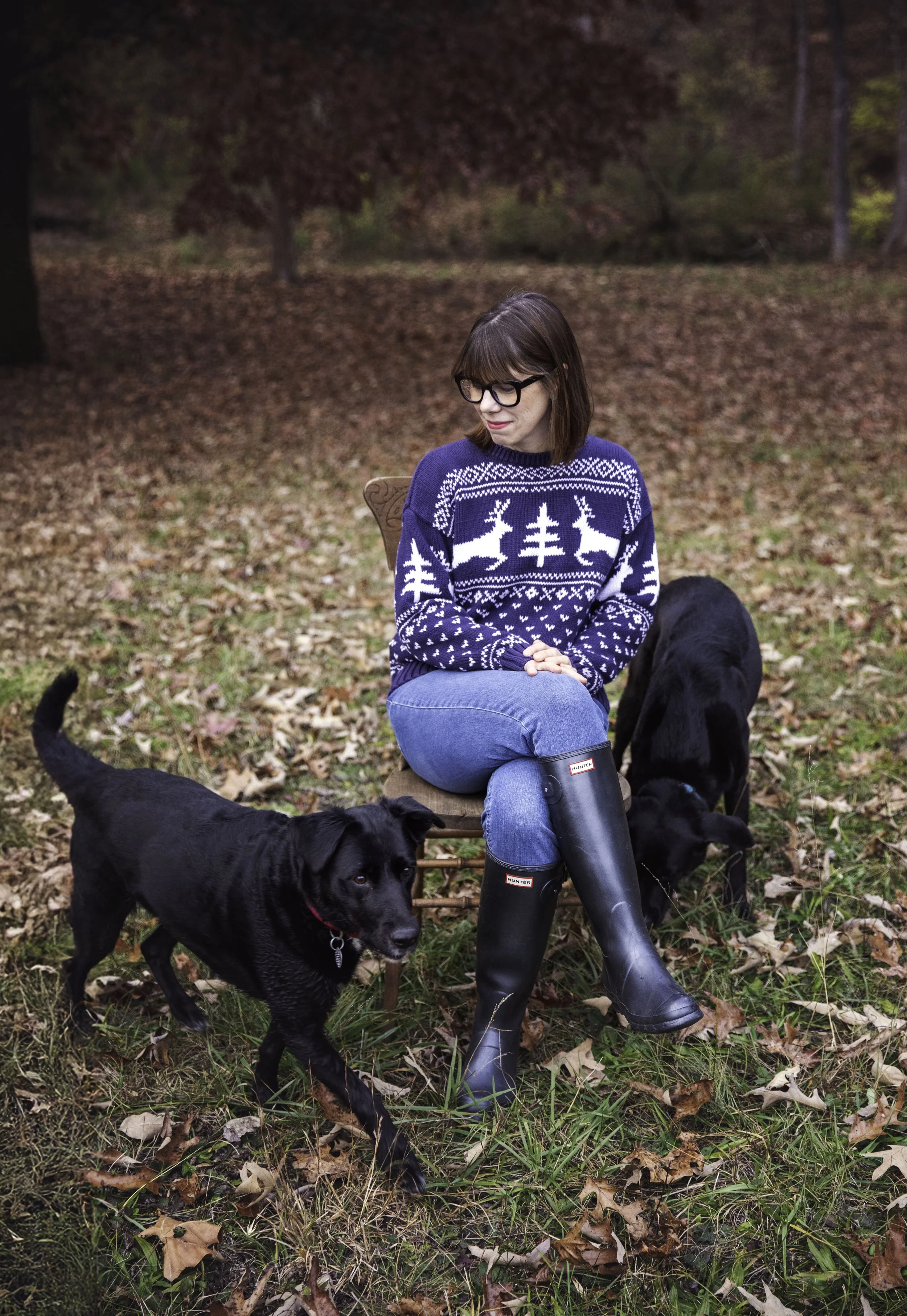 A woman with brown hair and glasses sitting outside in a wooded area during autumn, wearing a navy blue sweater with white winter patterns and black rubber boots, surrounded by three black dogs.
