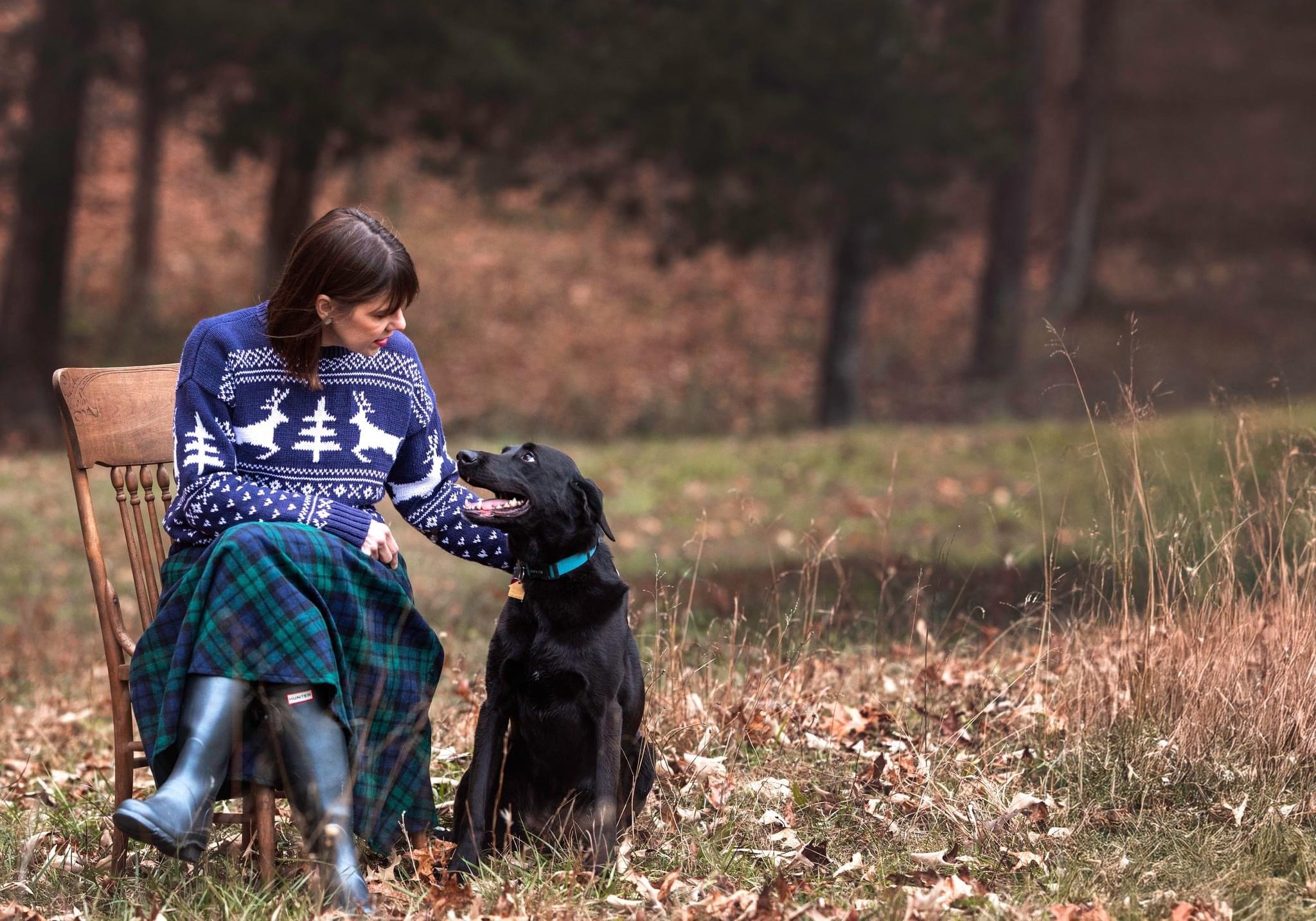 A woman in a blue Christmas sweater with white reindeer and trees, sitting on a wooden chair outdoors, looking down at a black dog sitting next to her, surrounded by fallen autumn leaves in a wooded area.