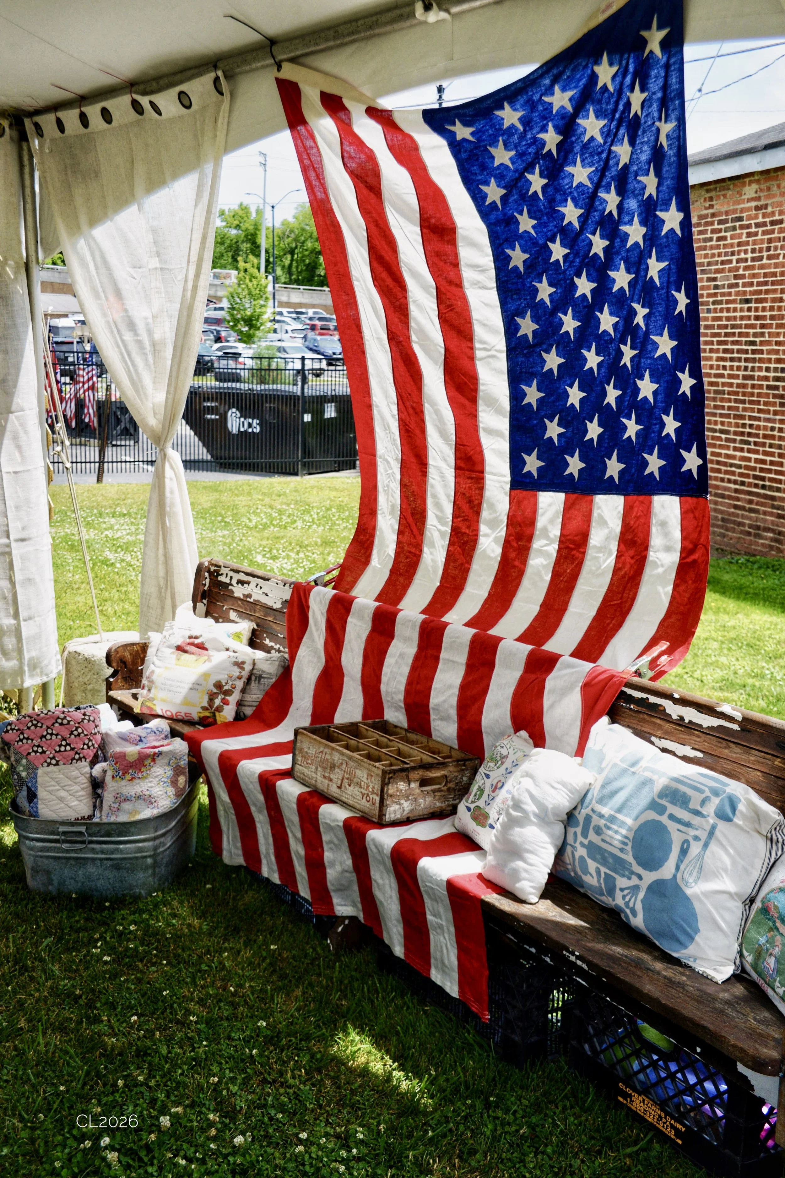 An American flag hangs inside a tent or booth at an outdoor event, with vintage and handmade items, quilts, pillows, and decorative pieces displayed on a wooden bench and nearby containers.