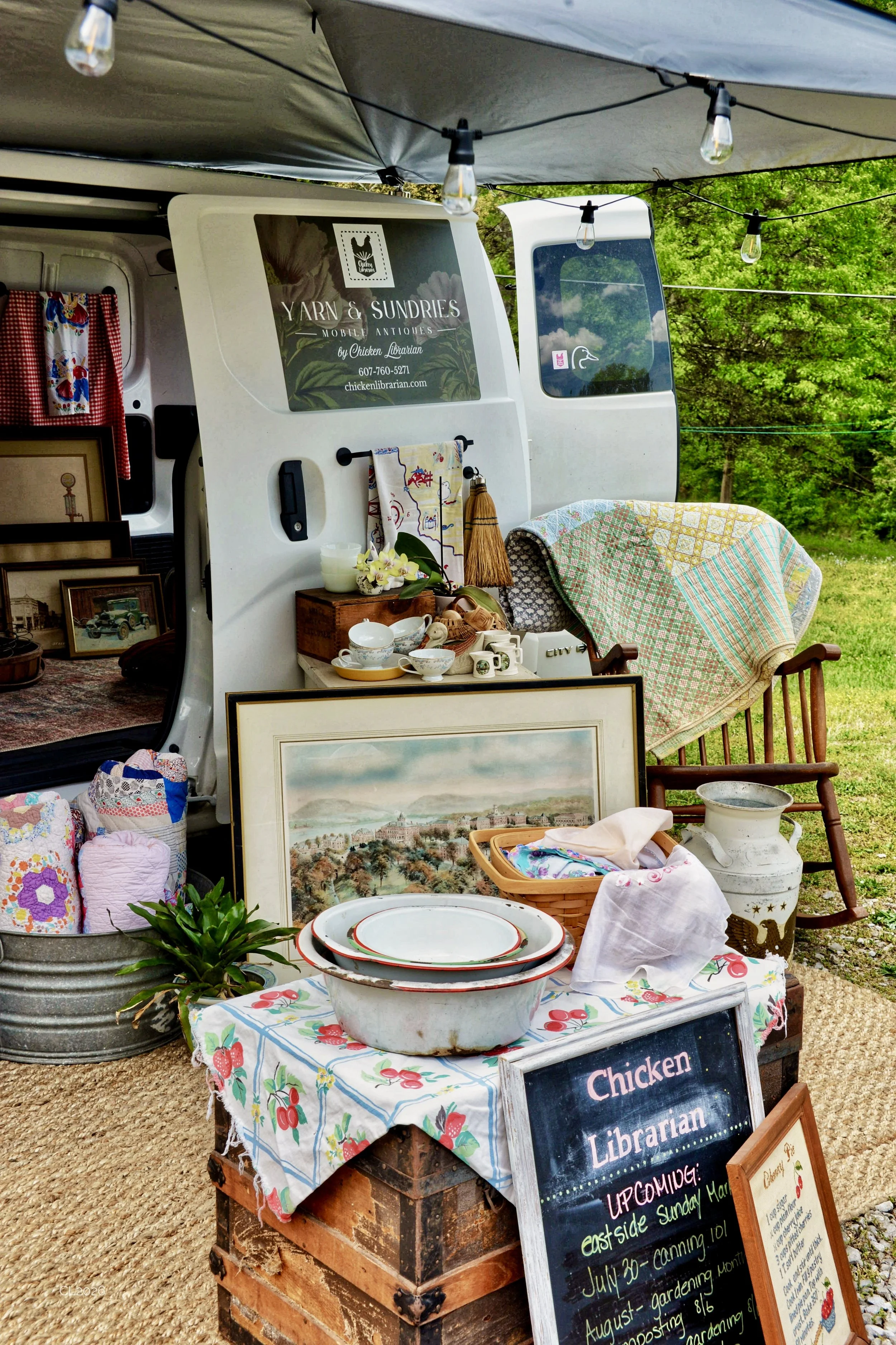 Outdoor yard sale with furniture, decorative items, and kitchenware displayed on tables and in containers, with a sign promoting chicken librarian antiques and upcoming events.