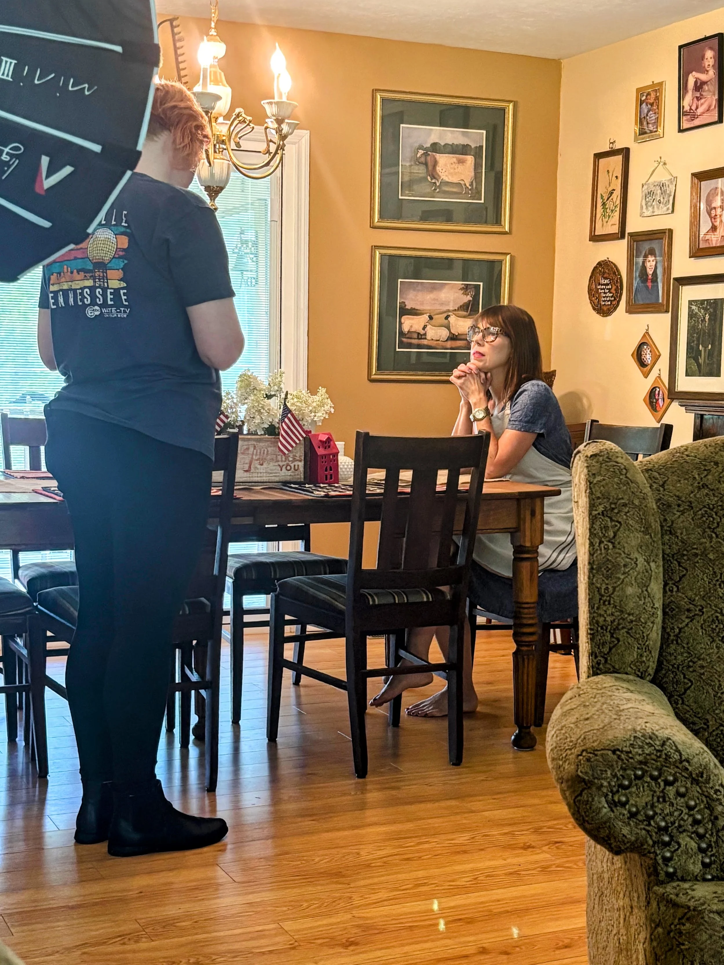A woman with glasses and dark hair seated at a dining table with her hands clasped, looking up at a person standing nearby. The standing person has red hair, wears a dark t-shirt and black shoes. The room has yellow walls decorated with framed pictur