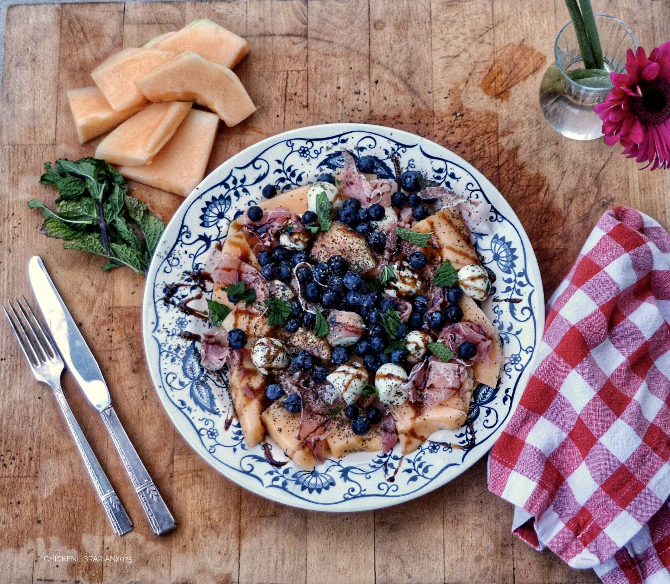 A round serving platter with blue floral design containing slices of melon and a salad with blueberries, sliced meats, and cheese, garnished with mint leaves. To the left of the platter, there is a bunch of mint leaves, and to the right, a pink flower in a small glass vase. A red and white checkered napkin is on the lower right corner and a fork and knife are on the left side of the image. The background is a wooden table.