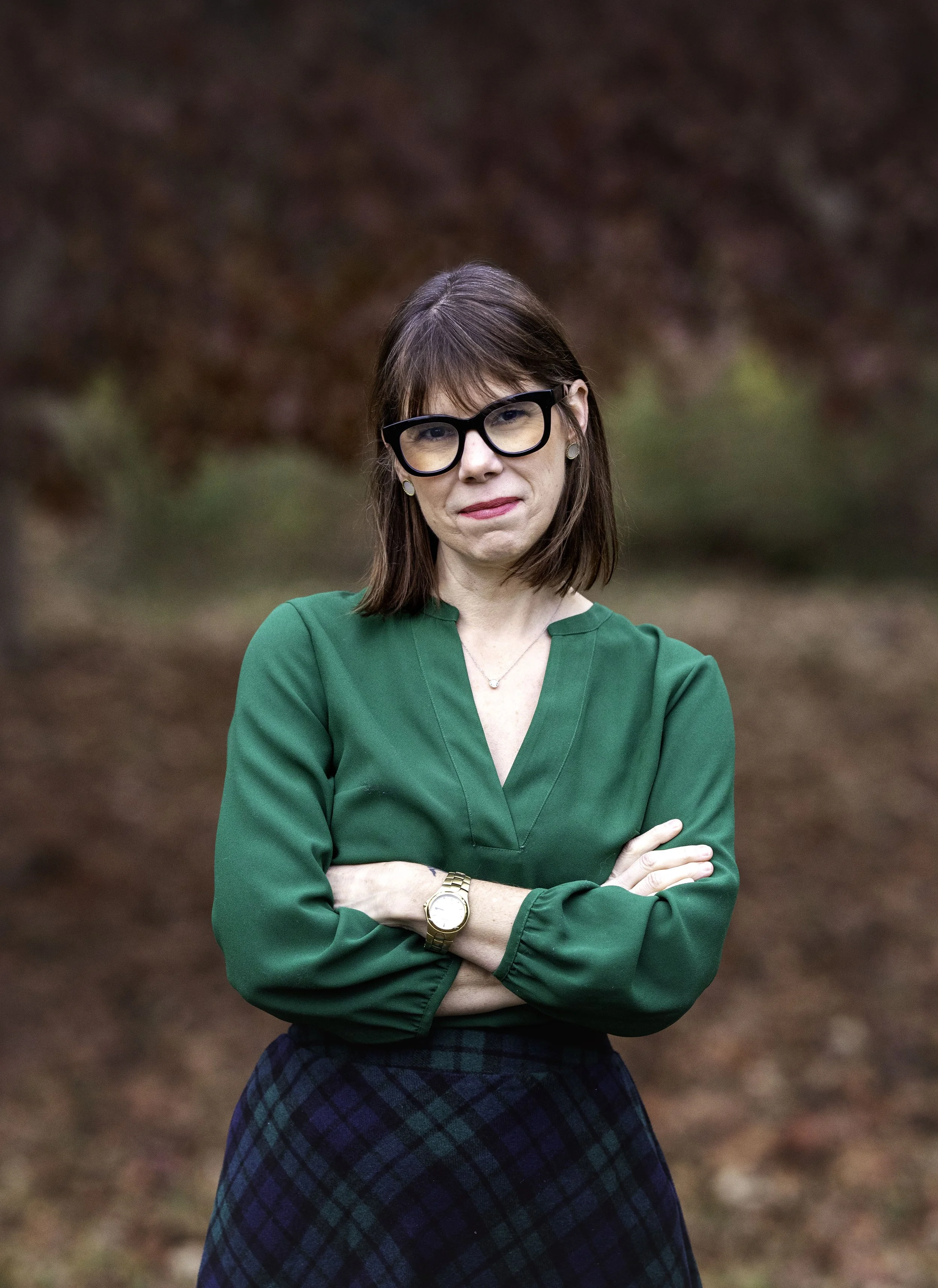 A woman with shoulder-length brown hair wearing black-rimmed glasses, a green blouse, and a plaid skirt standing outdoors with arms crossed, in front of blurred fall foliage.