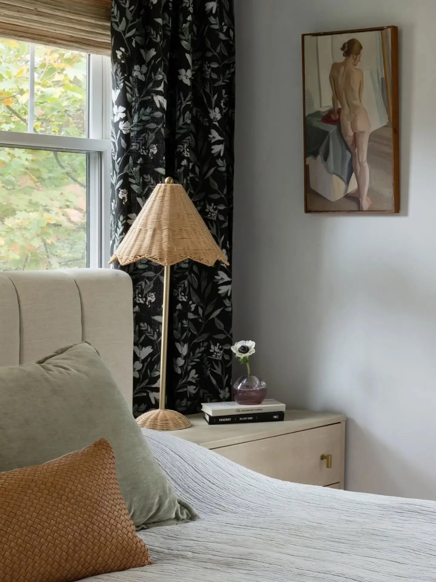 This bedroom strikes a balance between calm and contrast, with black drapery adding an unexpected twist. Natural elements, including woven Roman shades, a rattan lampshade, and a leather accent pillow, bring warmth, texture, and layers of interest to
