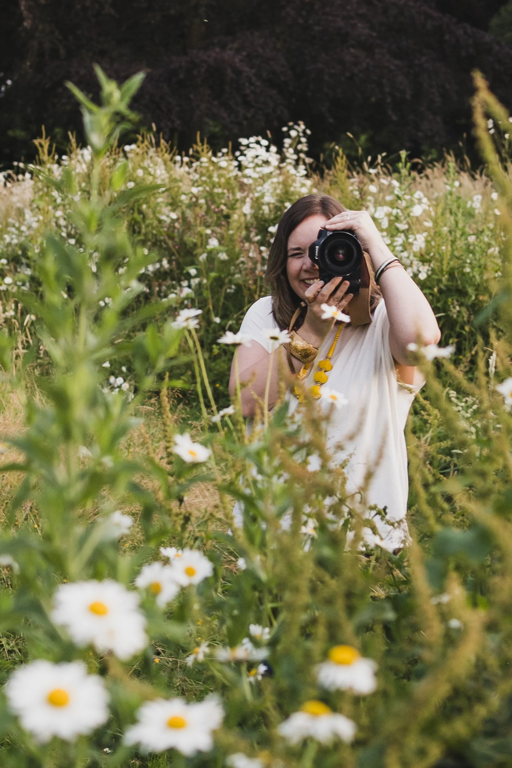 A lady sits in a daisy filled field and holds her camera