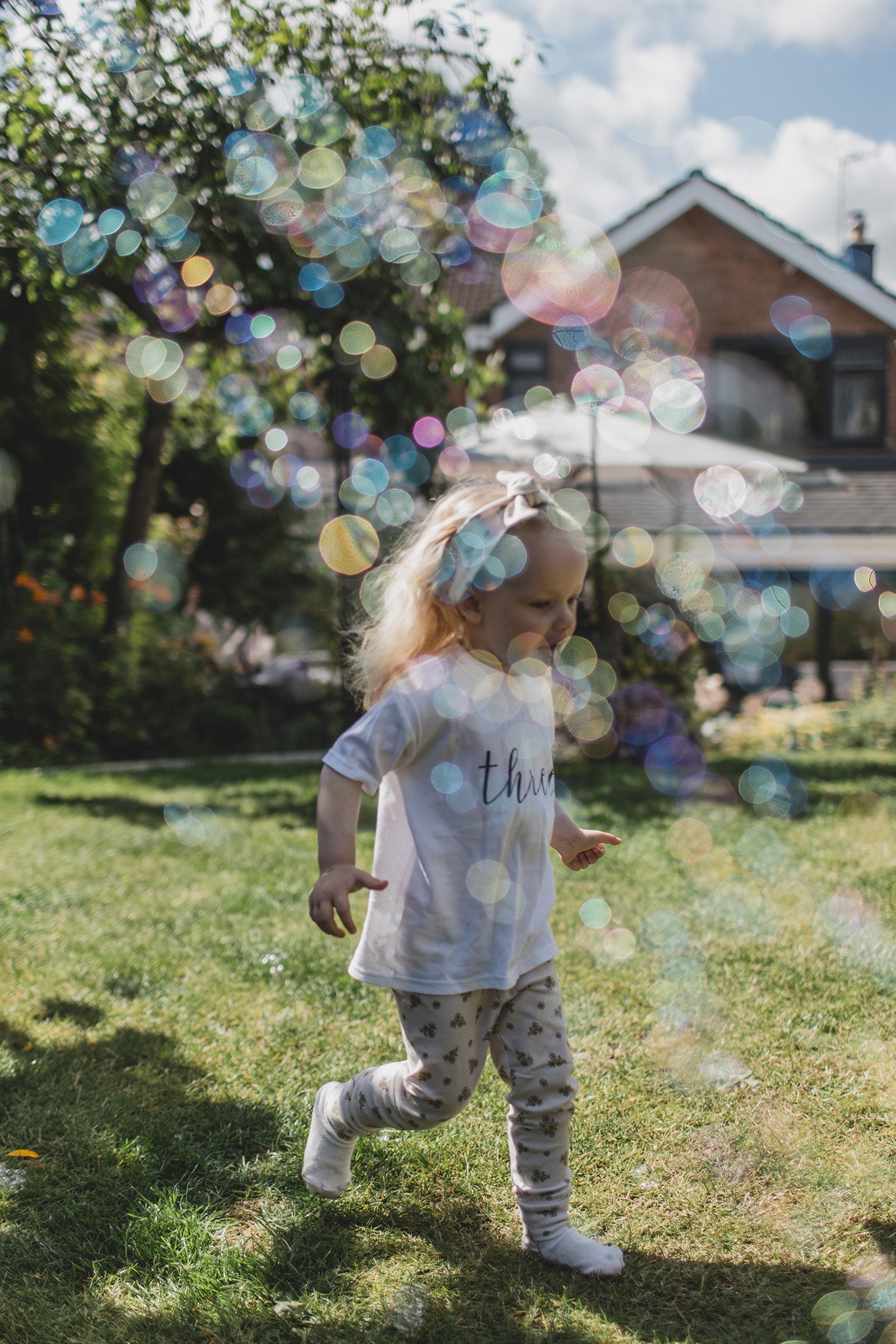 A child playing outside on grass with colorful bubbles in the air, with a house and trees in the background.
