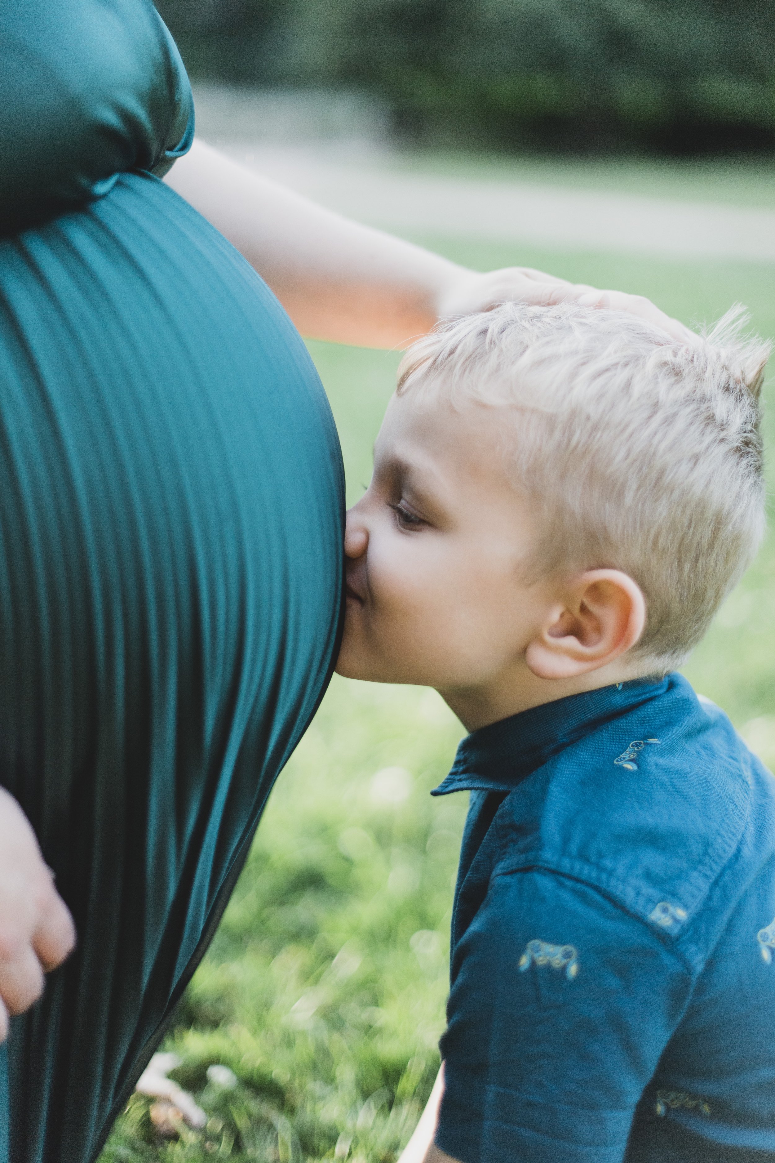 Young boy kissing a pregnant woman's belly outdoors.