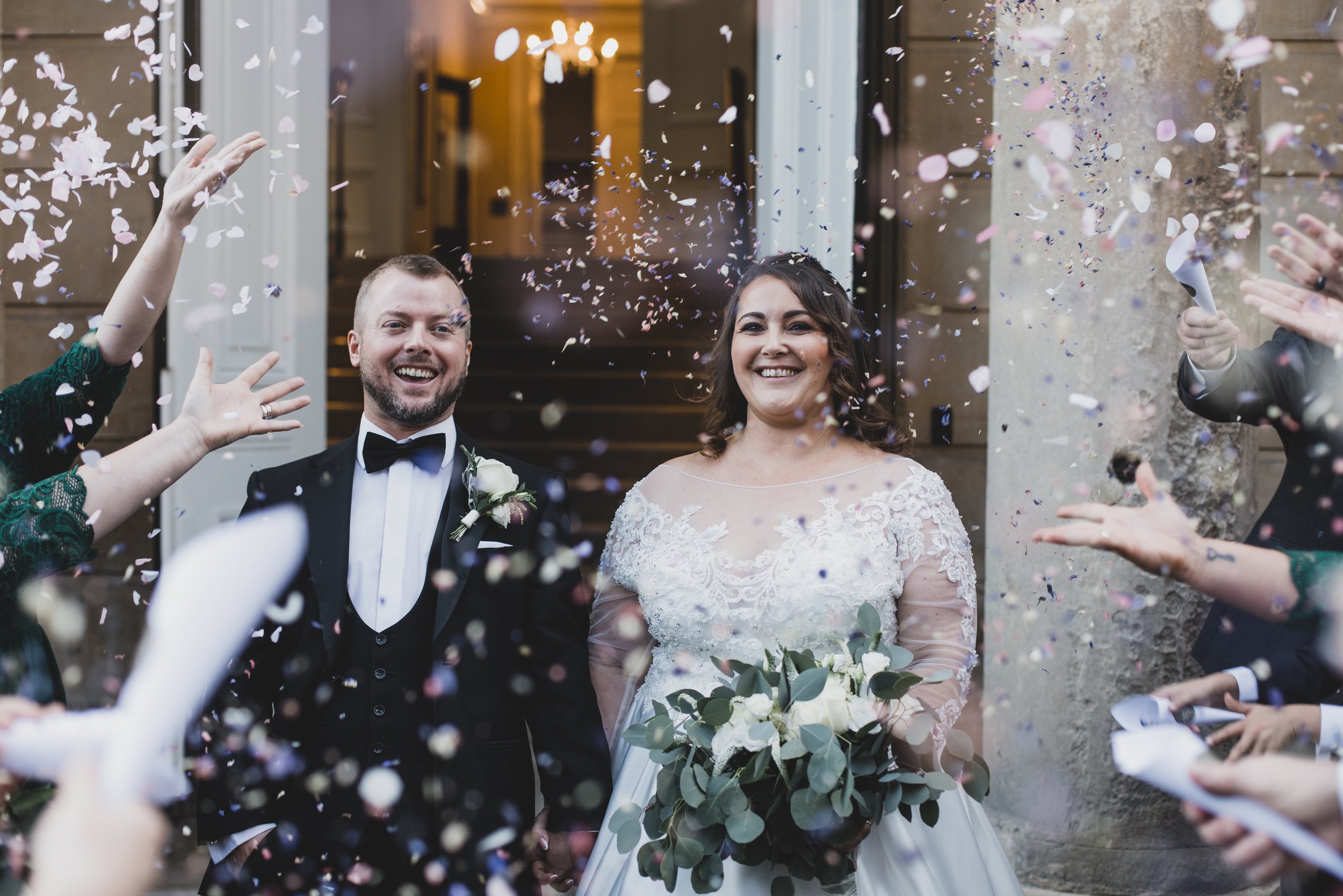 Bride and groom smiling while guests throw confetti at a wedding.
