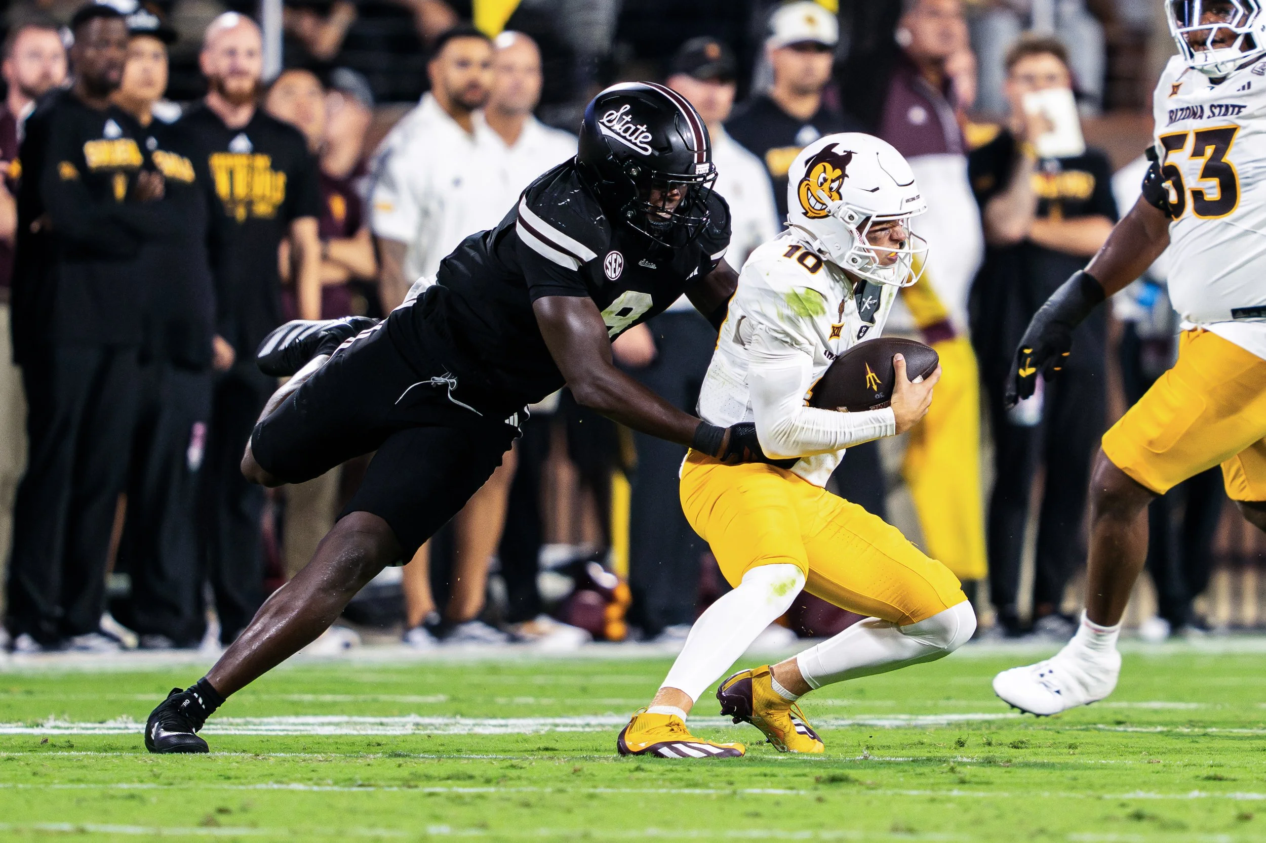 A football game with players from different teams, one in black uniform and another in white and yellow, during a play with a crowd of spectators in the background.
