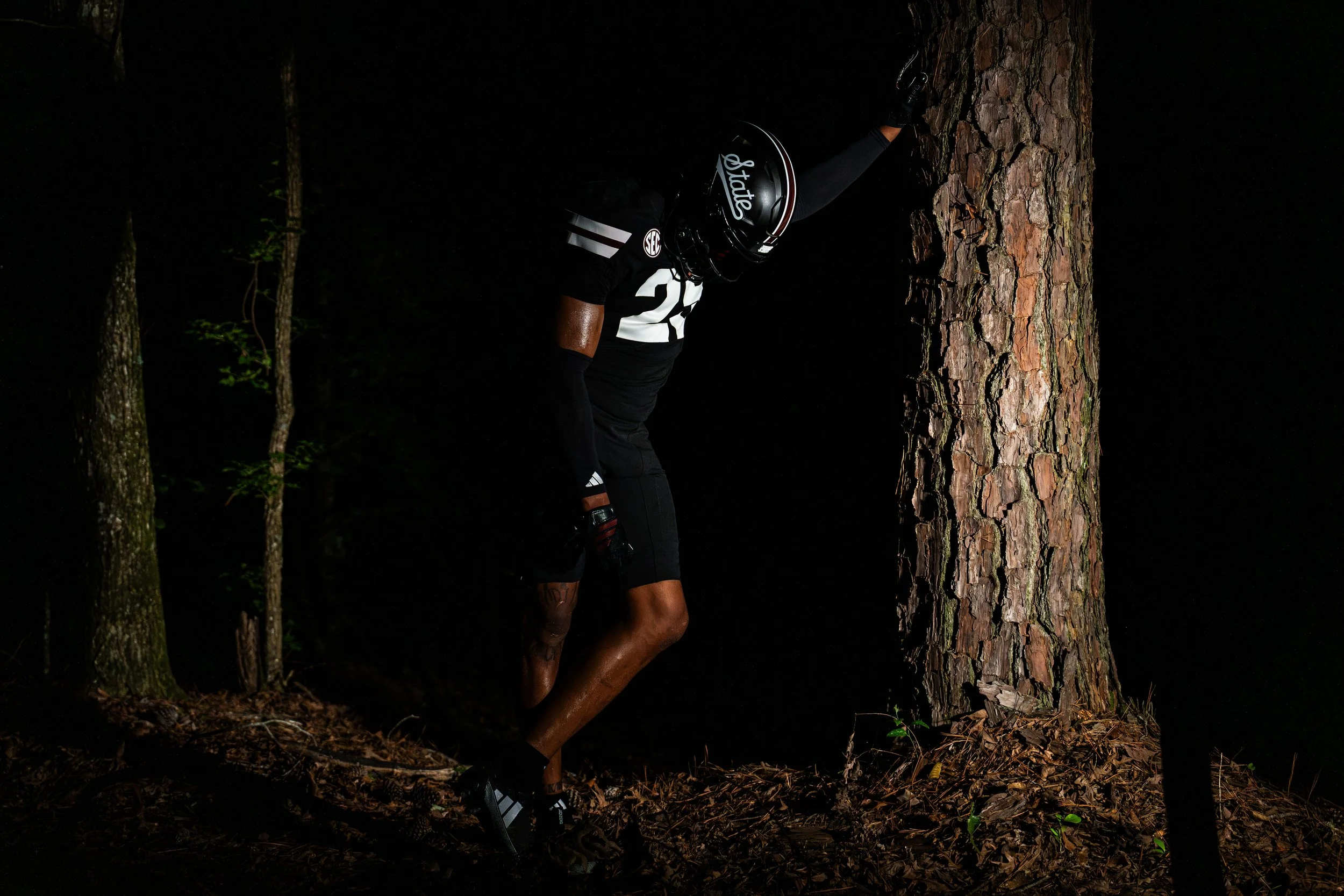 A football player wearing black uniform and helmet leaning against a tree in a dark forest at night.