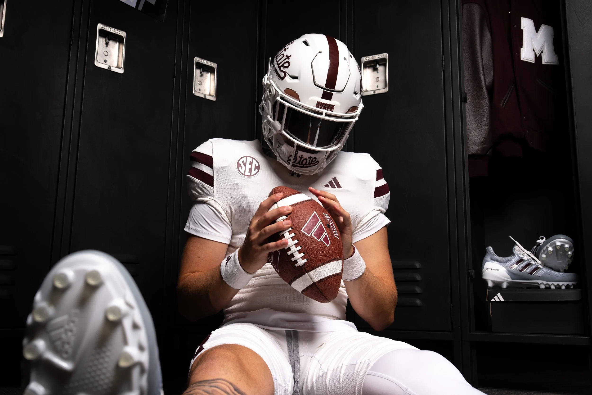 A football player in a white uniform and helmet sitting in a locker room, holding a football, with lockers and athletic shoes in the background.