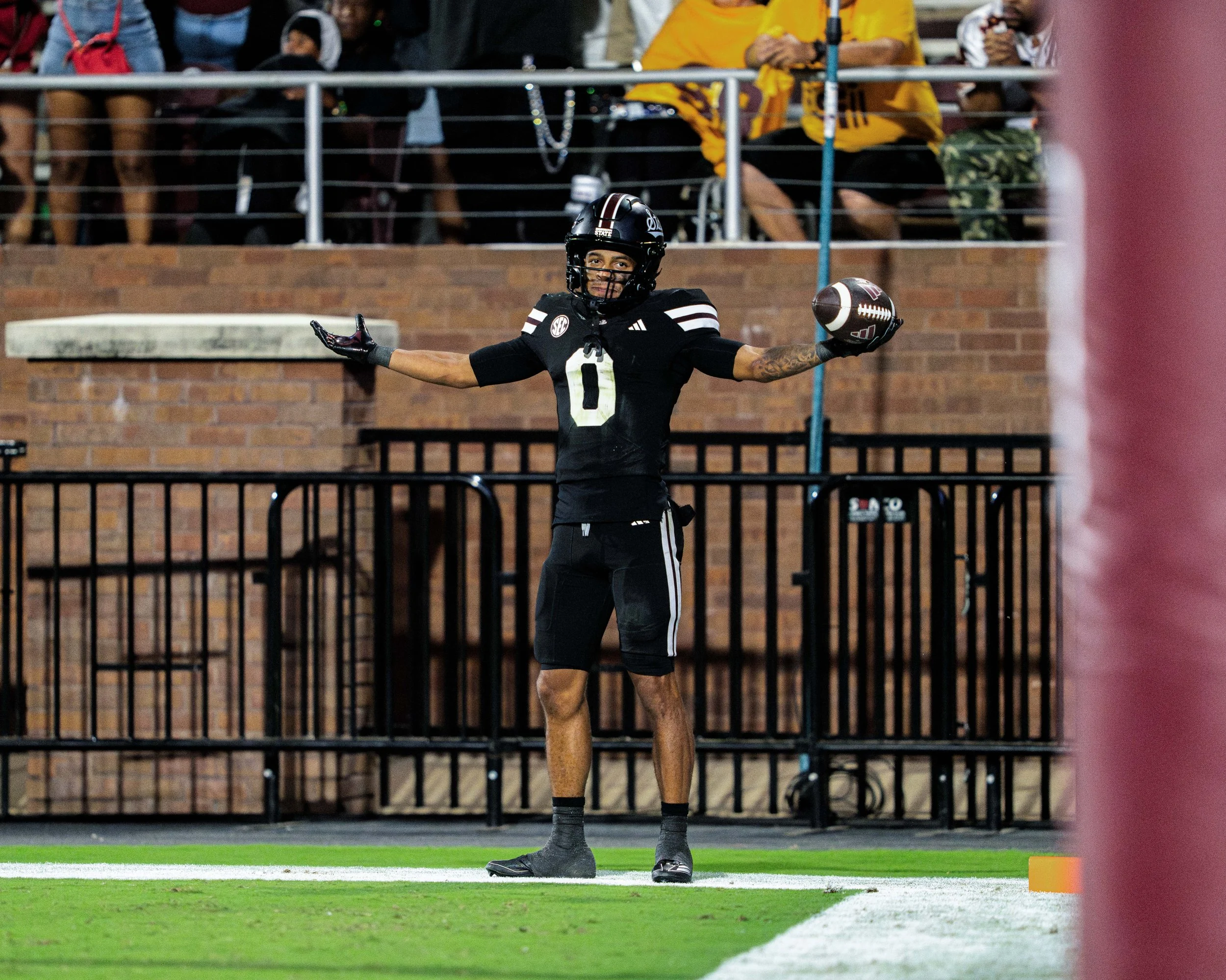 A football player holding a football in one hand on the field during a game, with spectators in the background.