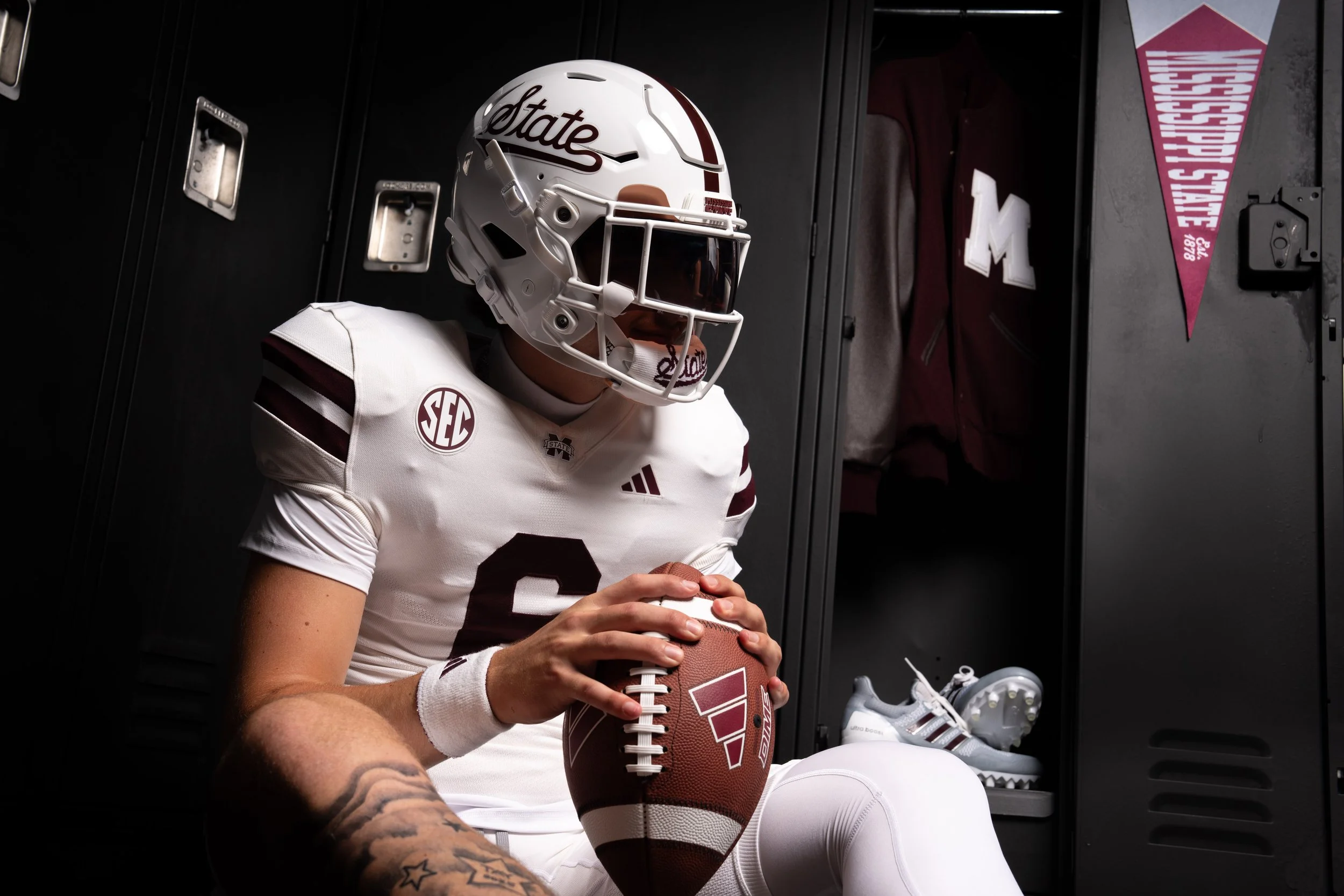 A football player wearing a white uniform with maroon accents, a white helmet with 'State' written on it, sitting in a locker room, holding a football, with lockers and a maroon letterman jacket in the background.