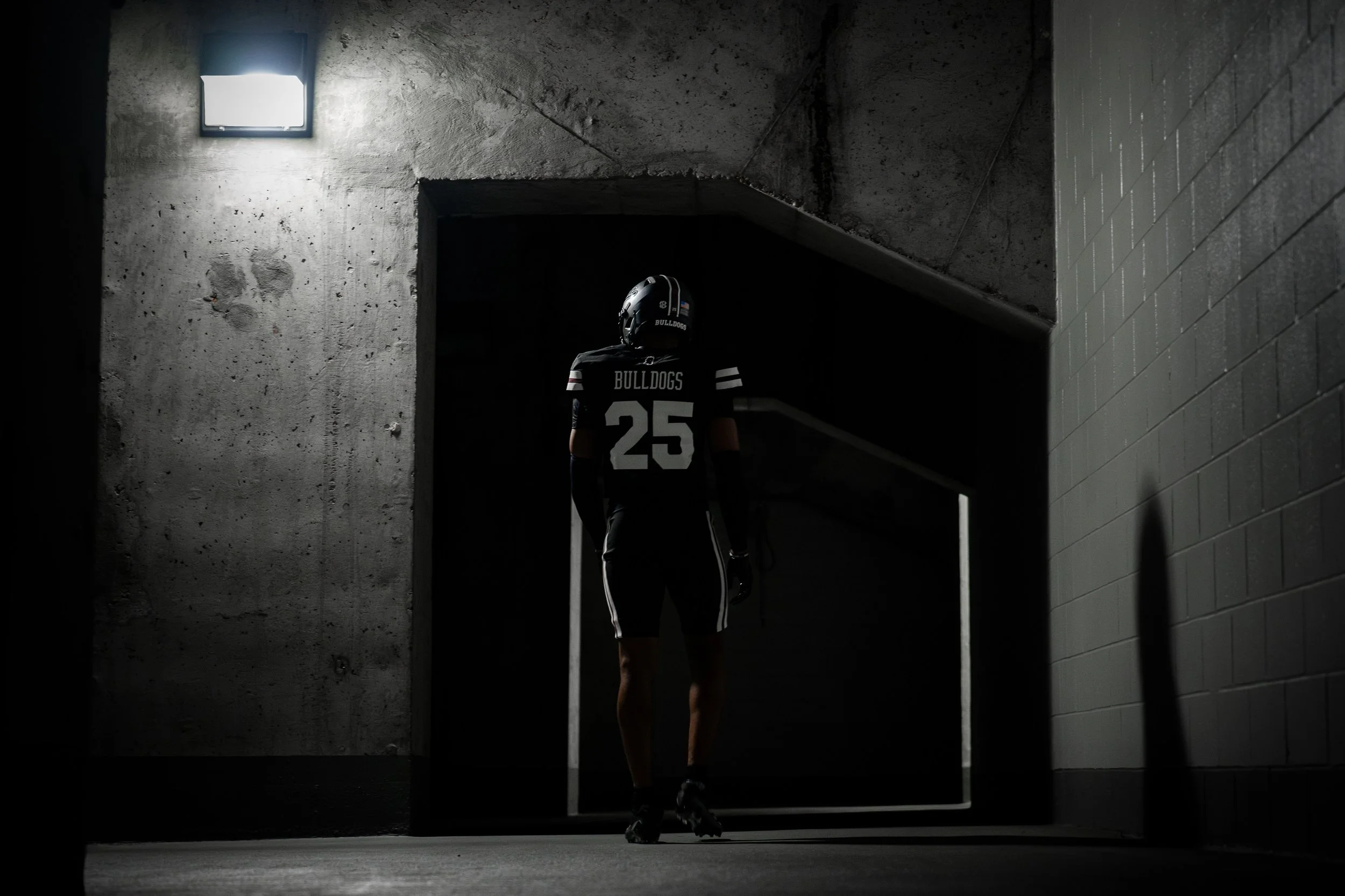 A football player in a black uniform with the number 25 and 'Bulldogs' written on the back, standing in an industrial-style hallway with concrete walls and a small window letting in light.