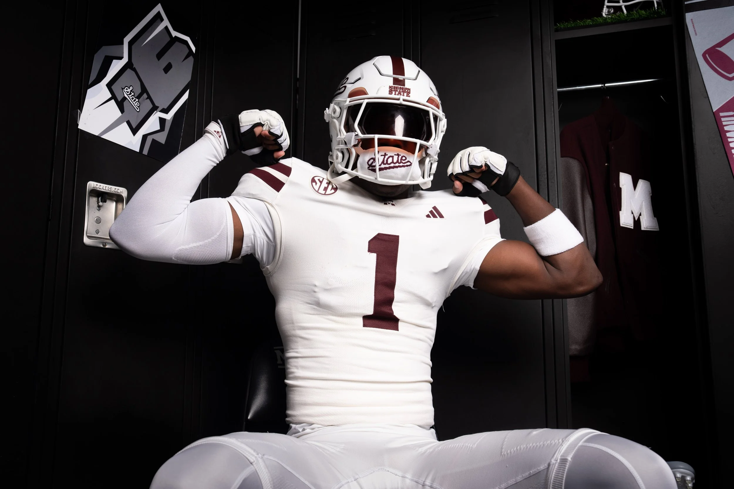 A person wearing a white football jersey with the number 1, a Mississippi State helmet and gloves, flexing their muscles in front of open lockers.