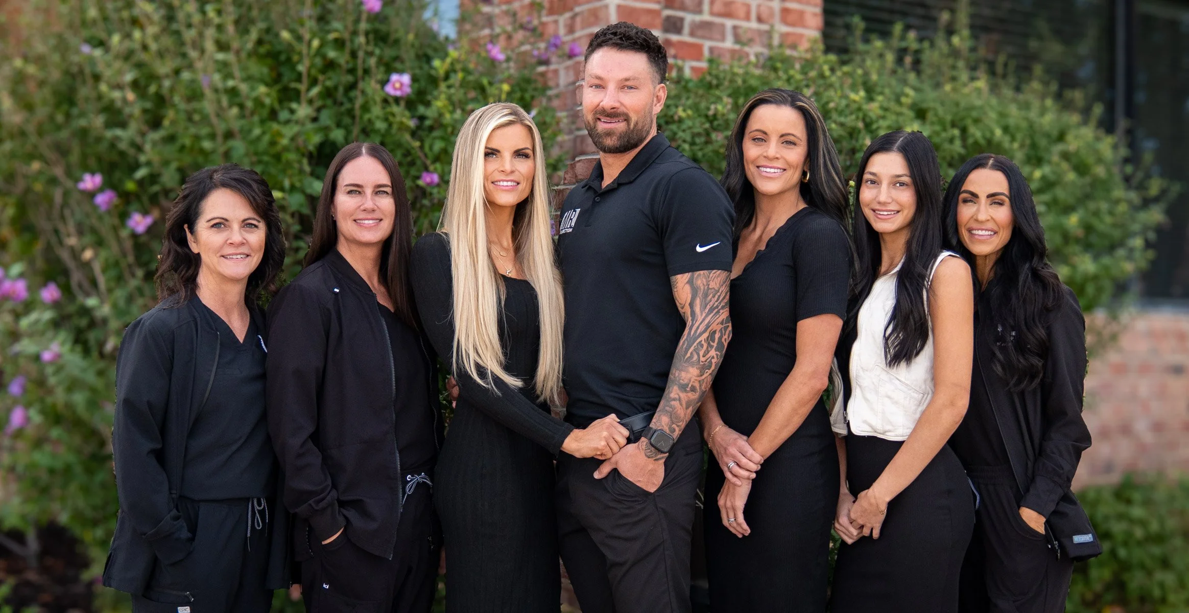 Group of seven people, six women and one man, standing outdoors in front of a shrub with purple flowers and a brick wall. They are dressed in casual and athletic clothing, smiling and posing for the photo.