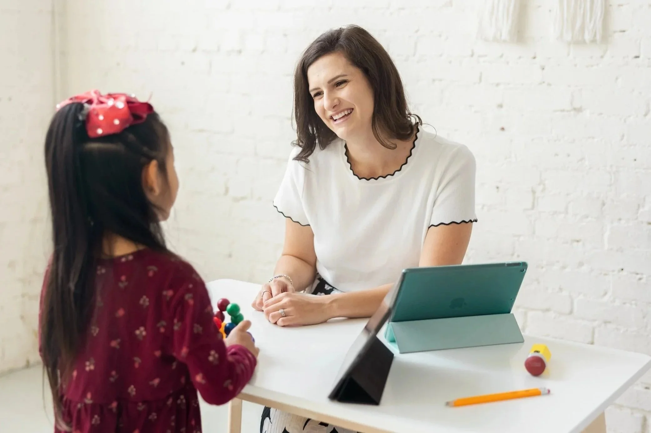 A woman with shoulder-length brown hair smiling at a young girl in a maroon sweater with a red bow in her hair. They are seated at a white table with tablets and toys, in a room with white brick walls.