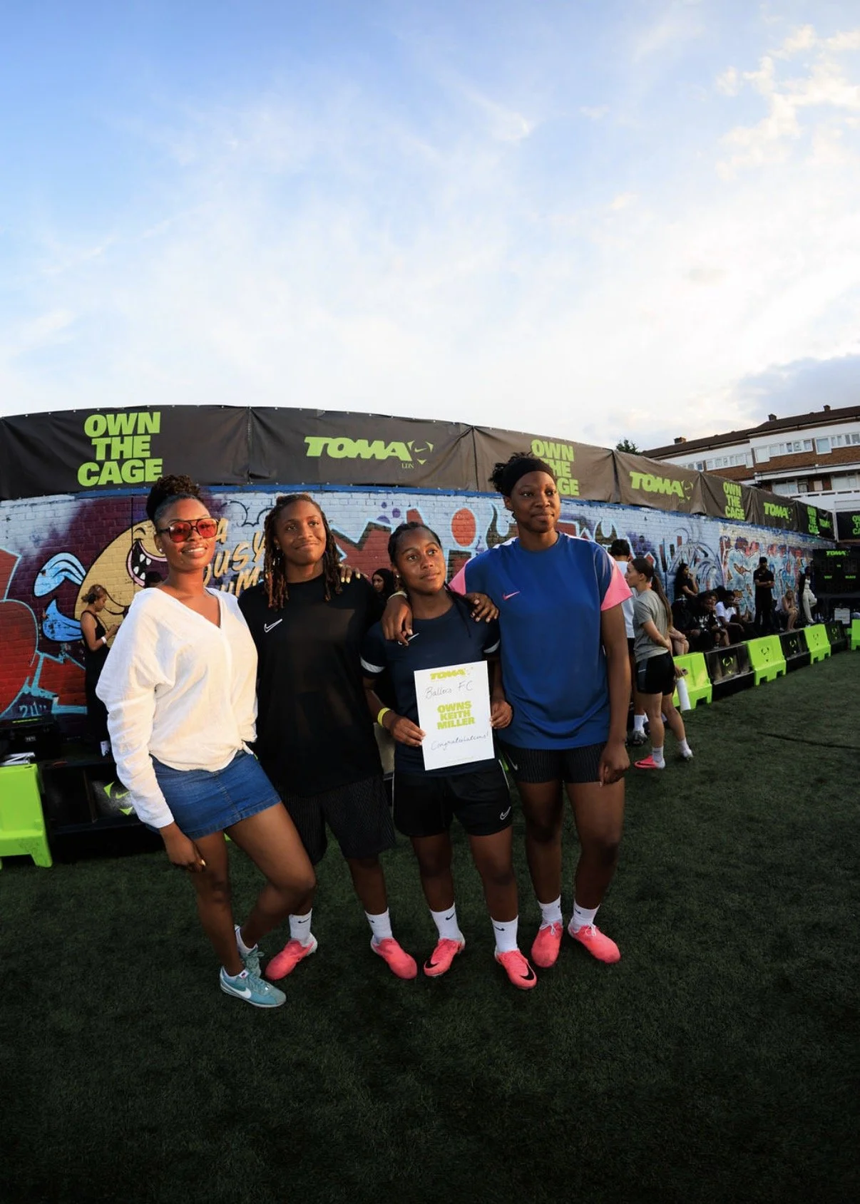 Four women standing together at an outdoor sports event, with a colorful graffiti wall in the background. One woman is holding a certificate that reads 'Congratulations,' and they are wearing athletic clothing.