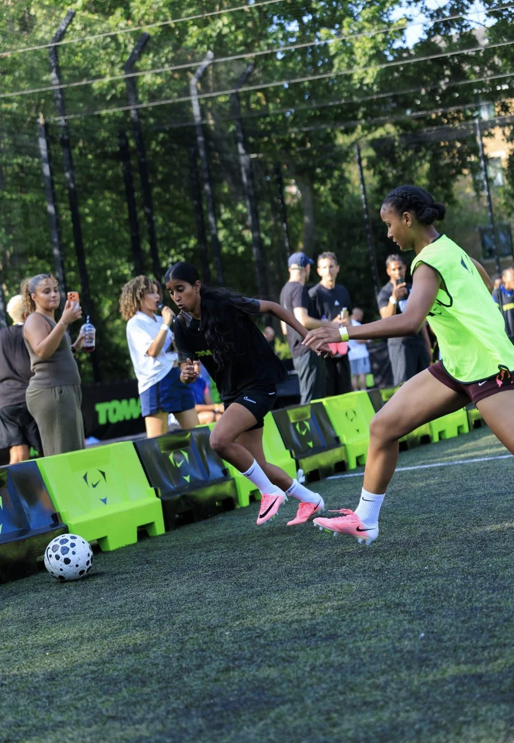 Two women playing soccer near the sideline on an outdoor field with green trees in the background, one in a black jersey and the other in a neon yellow jersey, while a group of spectators watches and takes photos.