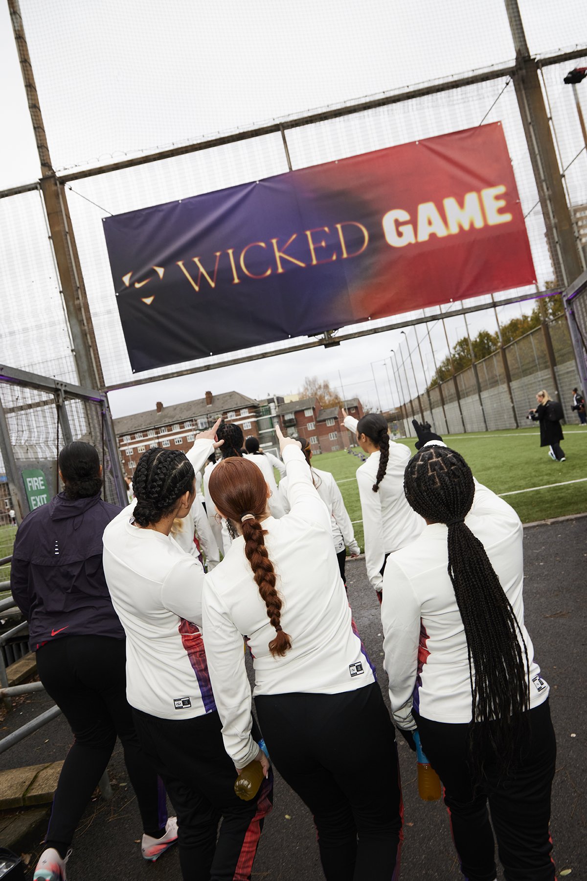 Women athletes in white jackets gather under a large sign that reads 'Wicked Game' at a sports field, with some pointing and holding drinks, and a fire exit sign visible on the fence.