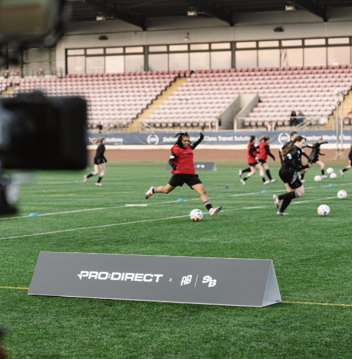 Girls playing soccer drills on a field with soccer balls, in a stadium with empty seats and a sign that reads "PRO:DIRECT".