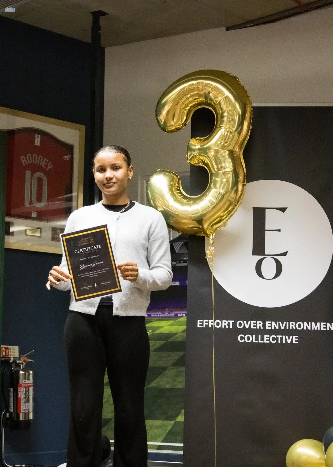 Young woman standing indoors, holding a certificate, with a large gold '3' balloon and an 'E3' banner promoting effort over environment collective in the background.