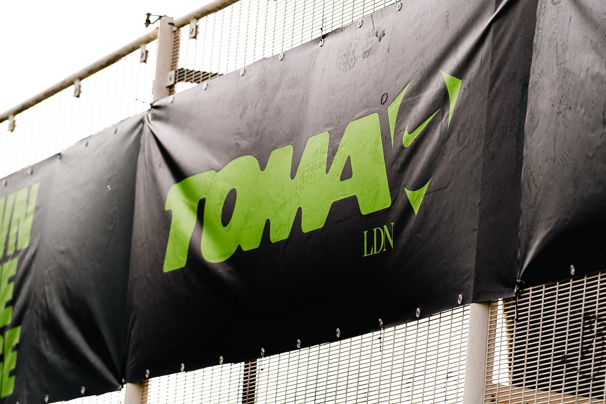 Close-up of a black advertising banner with green text, hanging on a metal fence. Part of a white wall and overhead light fixture are visible in the background.
