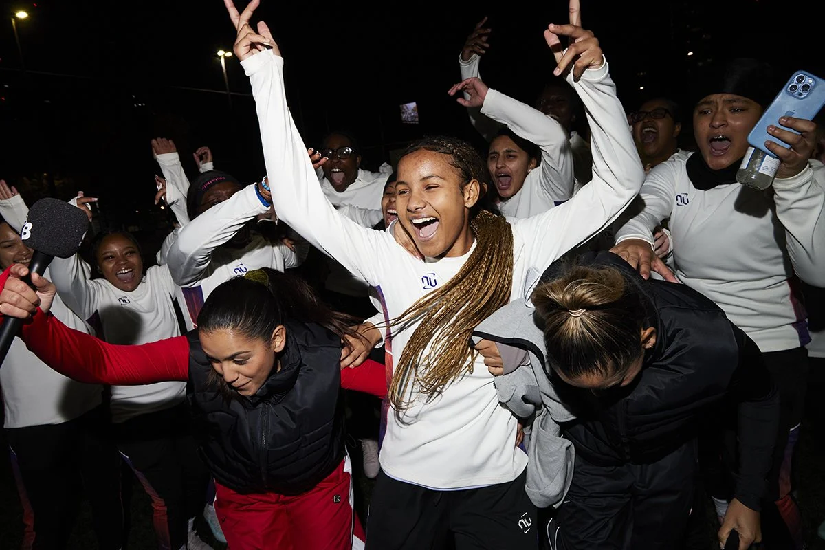 Group of women celebrating, some dancing and smiling, at night with dark background, wearing sportswear, one woman in the center with long braided hair and arms raised in victory.