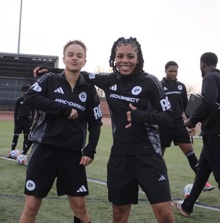 Two female soccer players in black athletic gear standing on a soccer field, smiling and posing for a photo, with other players and people in the background.
