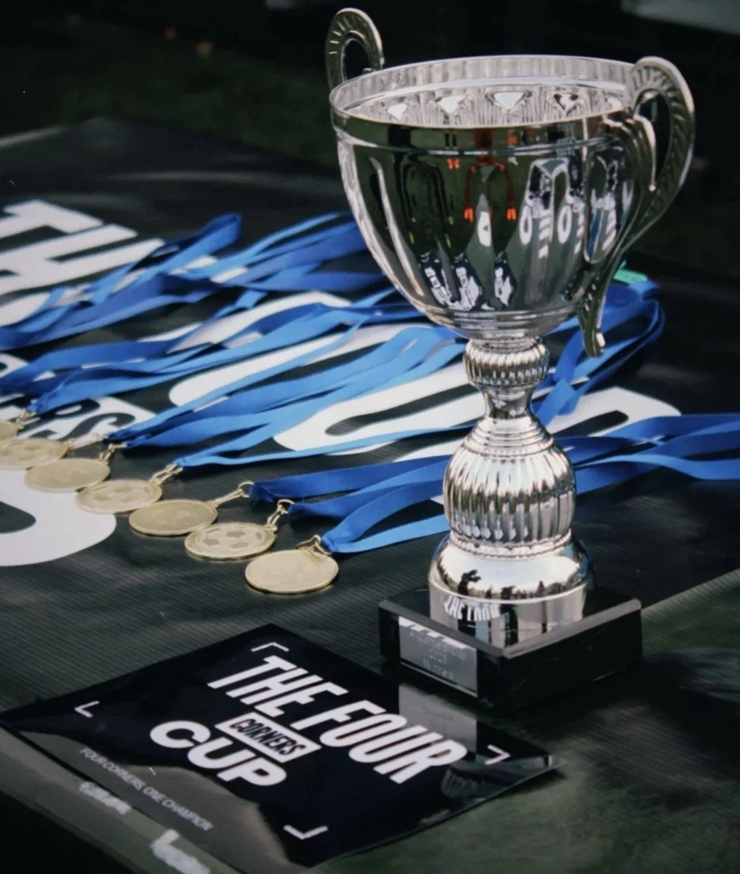 Silver trophy cup and several medals with blue ribbons laid out on a dark surface, and a black and white sign that reads 'THE CUP'.