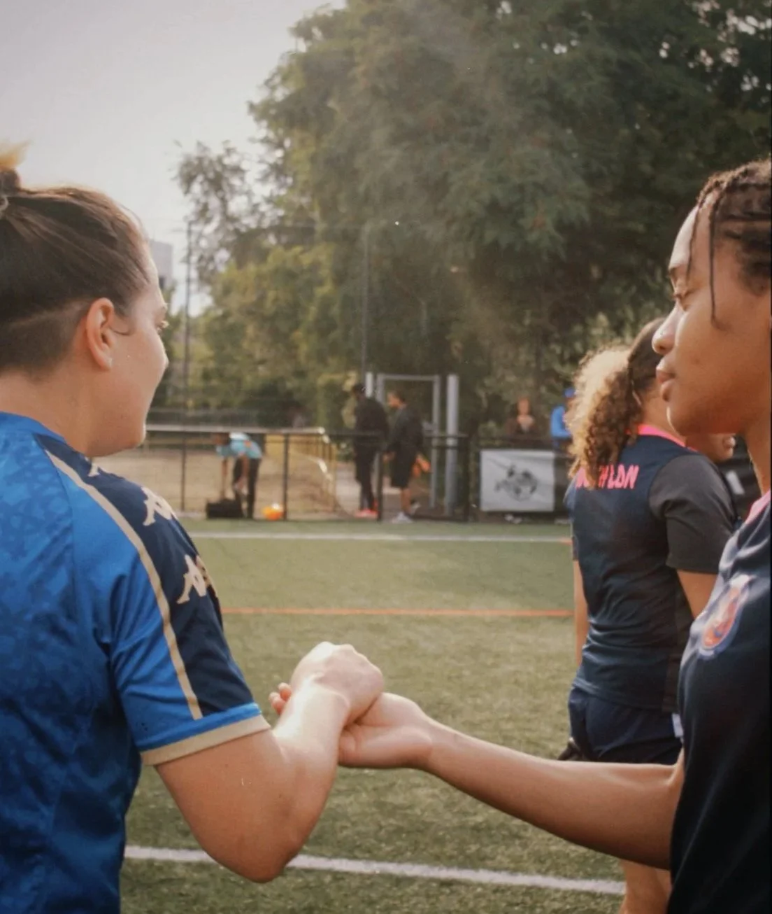 Two women on a sports field engaging in a handshake or fist bump, with others and a fence in the background, during daylight.