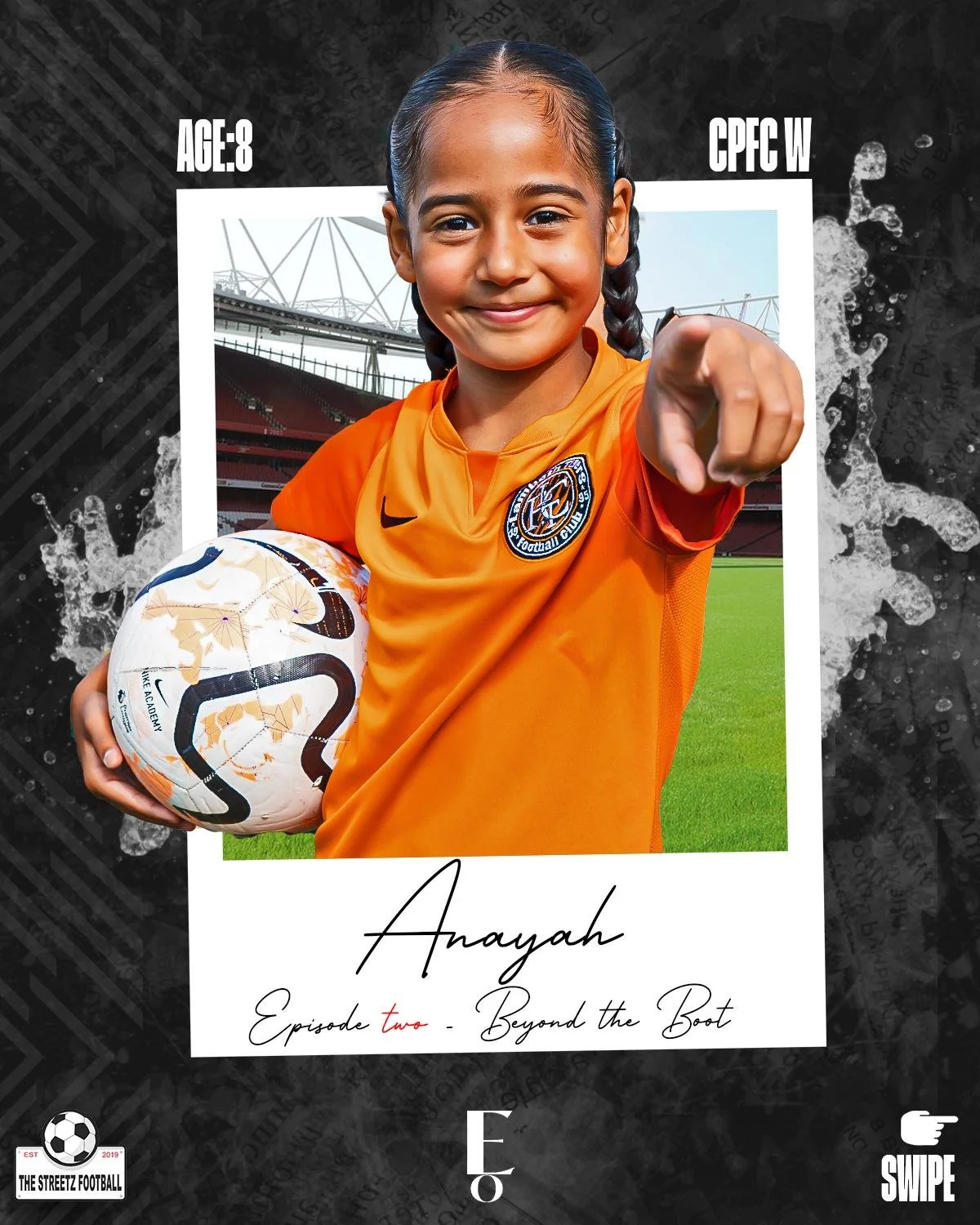 Young girl in orange soccer jersey holding a soccer ball points directly at the camera, standing on a soccer field with stadium in the background.