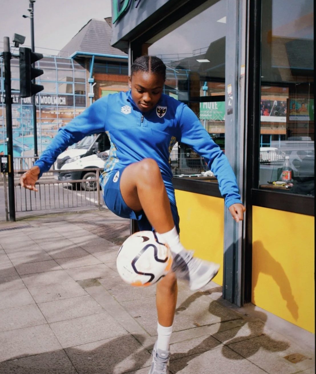 A young woman in a blue sports outfit is juggling a soccer ball on a sidewalk outside a store with large glass windows.