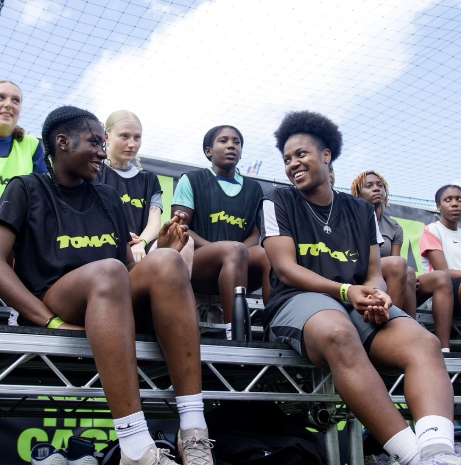 Group of female athletes sitting on bleachers inside a dome, wearing black TAWA shirts, some smiling and engaged in conversation.