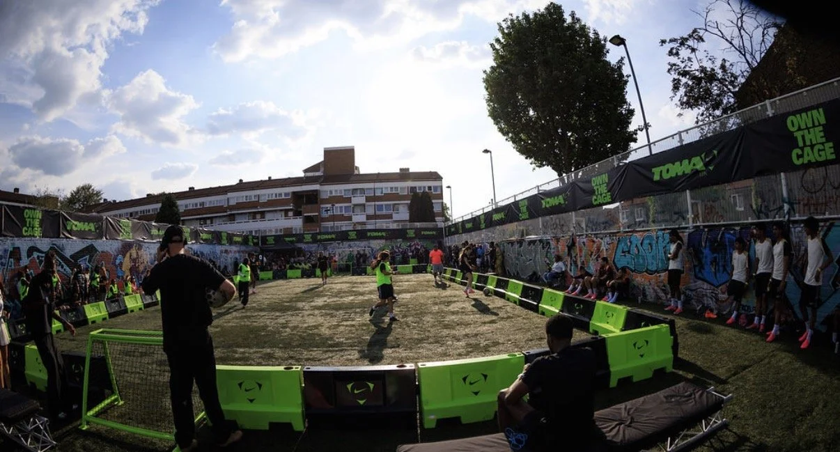 A soccer field surrounded by bright green barriers with people playing a match, spectators on the sidelines, graffiti on the walls, and a building with multiple floors in the background under a partly cloudy sky.