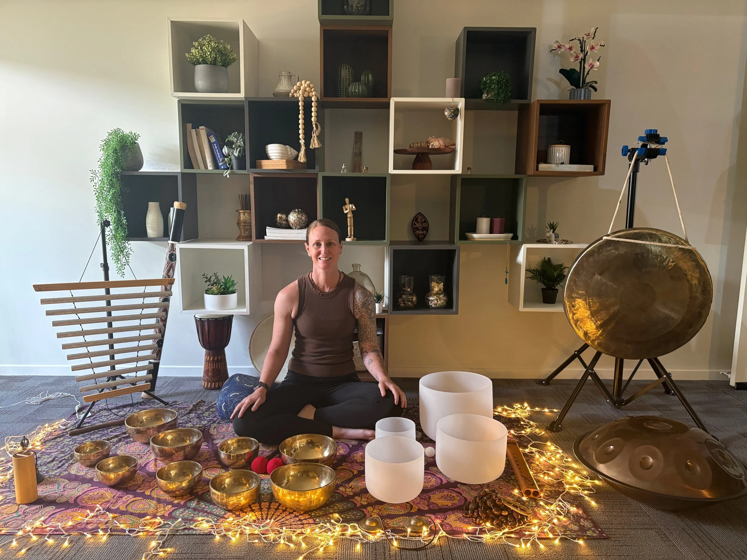 A woman sitting cross-legged on a decorative purple and pink patterned rug, surrounded by seven gold singing bowls, holding a wooden mallet. She is smiling and wearing a dark green polo shirt with a logo that says 'Botanica Massage'. There is a shelf with various decorative items and plants in the background.