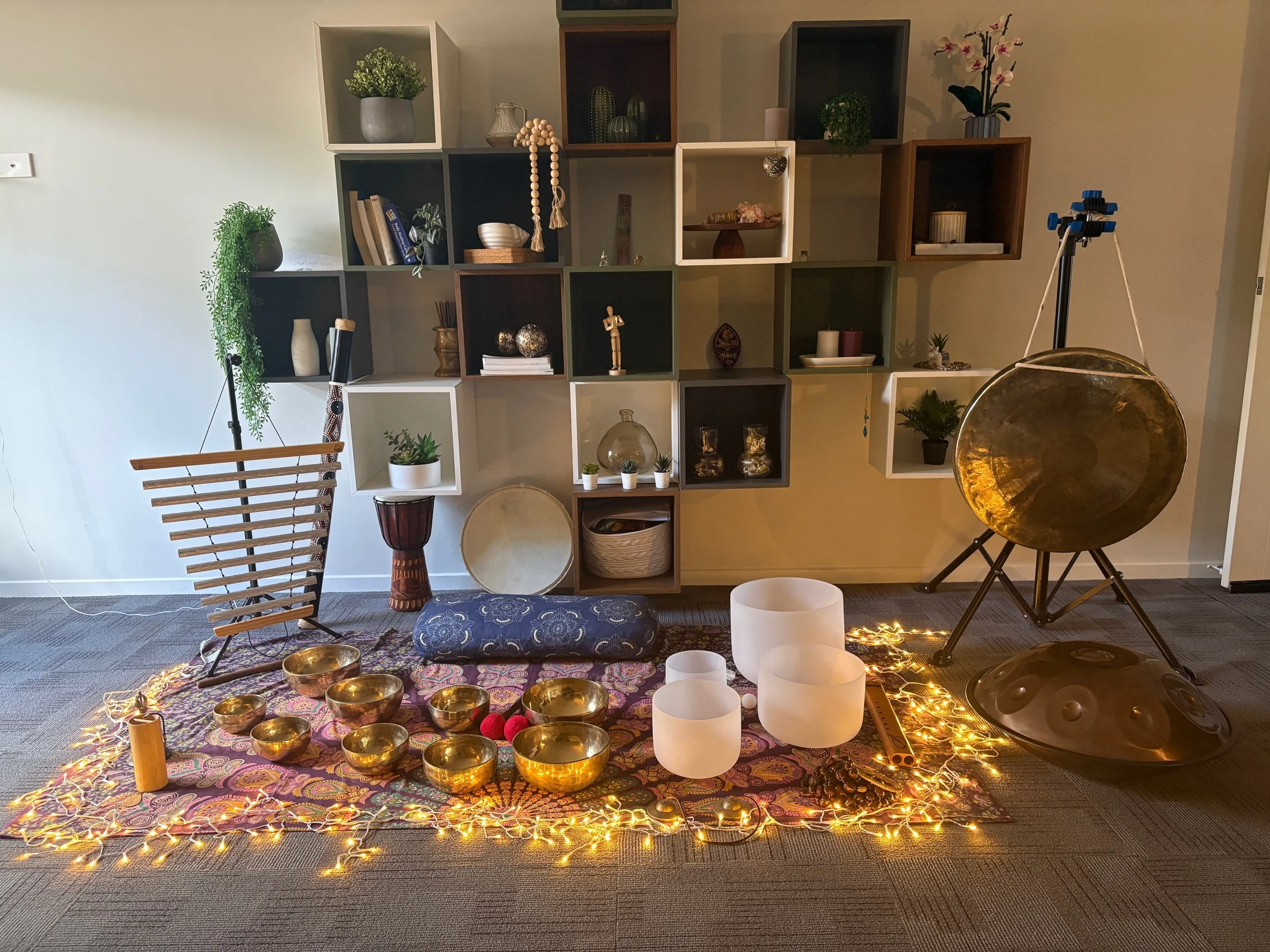 An setup for a sound therapy or meditation session with a colorful paisley patterned cloth covering the floor. There are seven metallic singing bowls of various sizes arranged in a semi-circle, two mallets with red felt balls, a blue cushion, a wooden stick, a colorful decorative rock, a painted rock, a wooden stick with a black knob, a rainbow-colored fabric-covered object, and a small rectangular cushion at the top of the setup.
