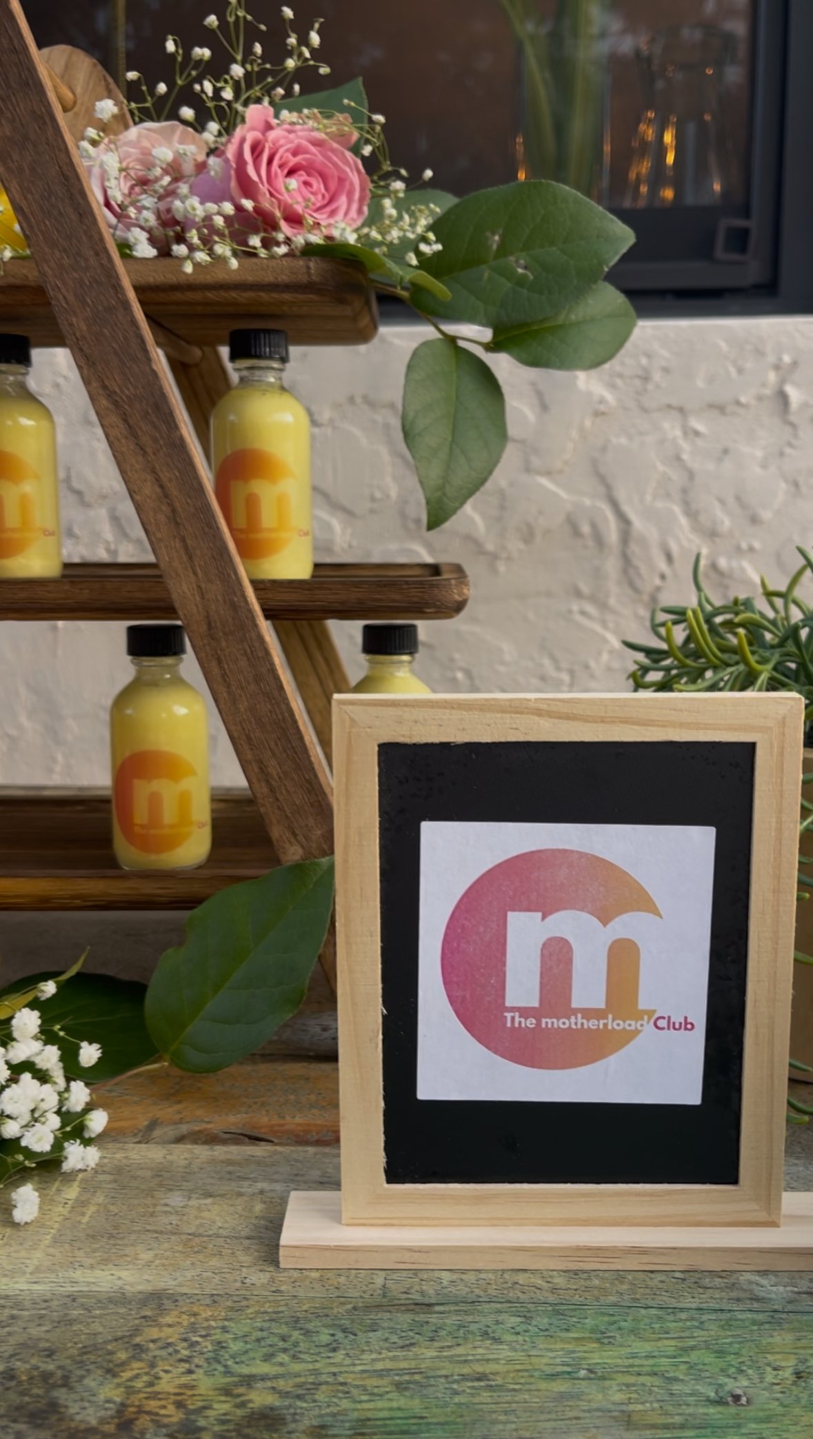 Decorative display with small yellow bottles, flowers, and a wooden sign with "The Motherload Club" logo.