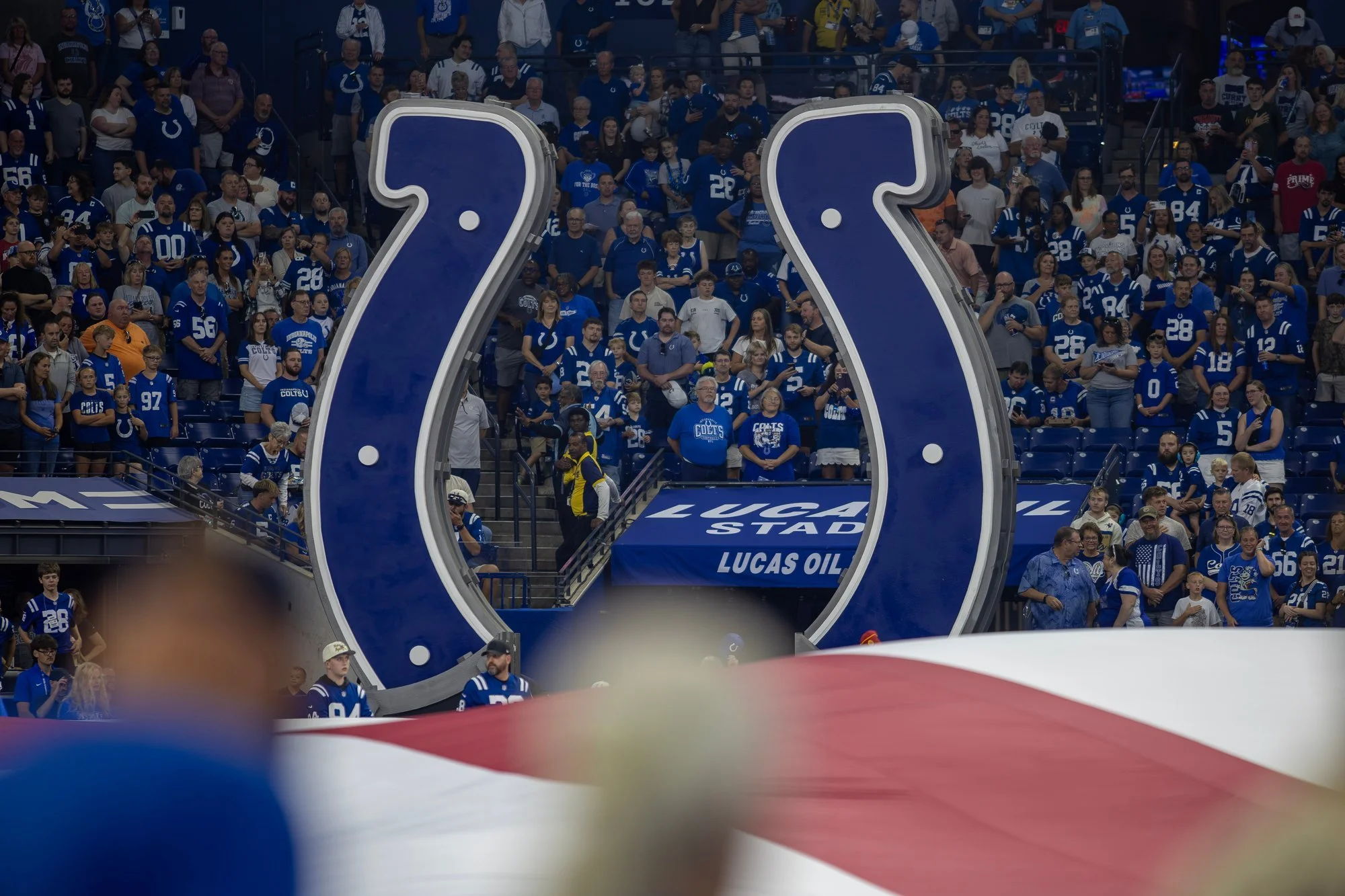 Large Colts horseshoe logo inside Lucas Oil Stadium with fans in the stands behind it.