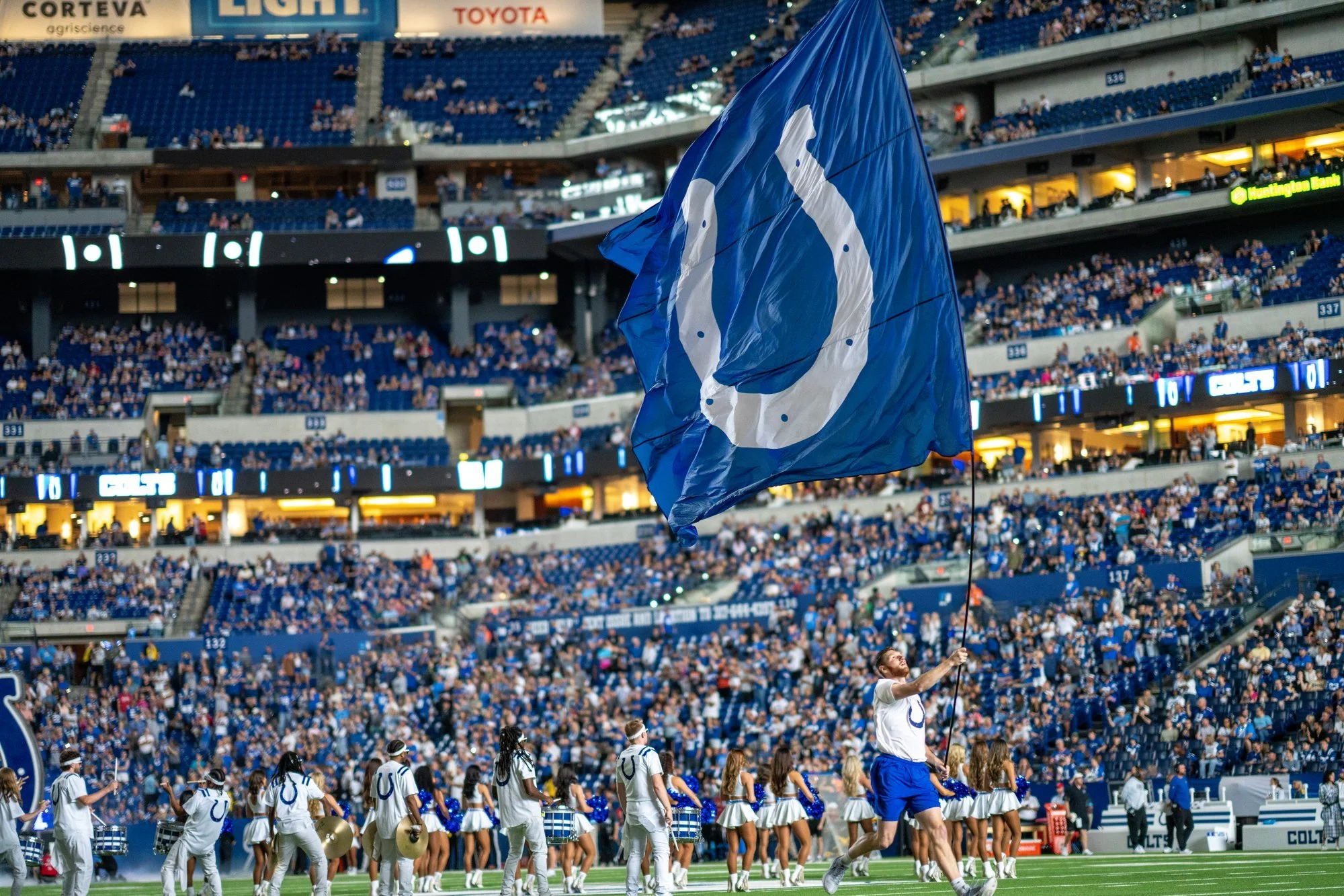 Colts cheerleaders on the field below a giant horseshoe flag in a packed Lucas Oil Stadium.