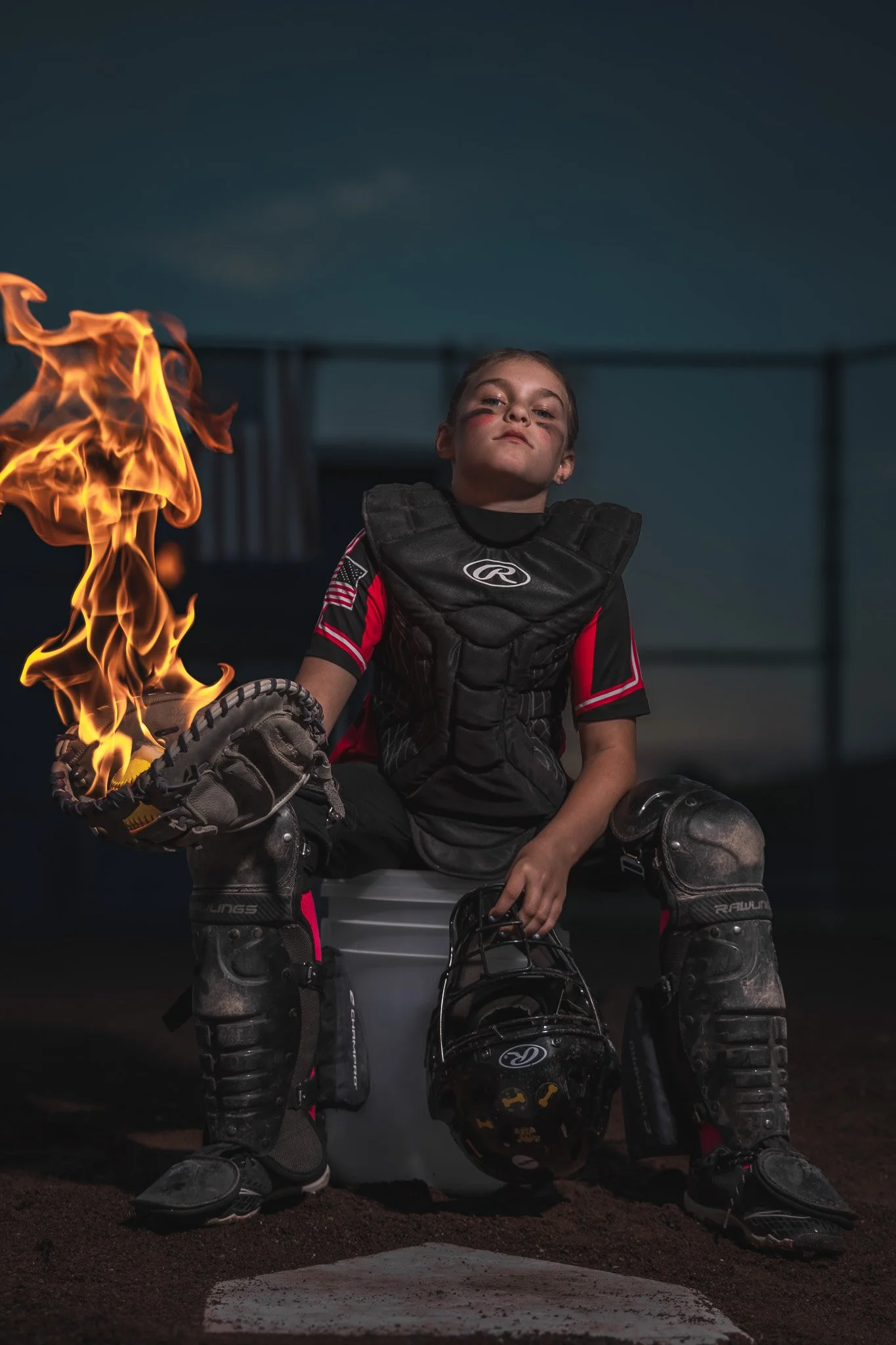 Young softball catcher in black chest protector and leg guards sitting on a bucket at home plate at dusk, holding a catcher's mitt that is on fire