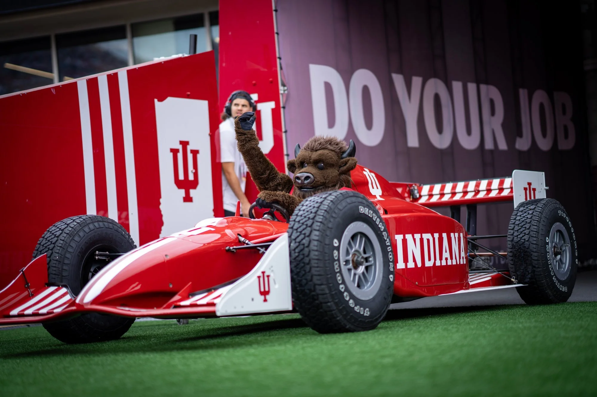 Costumed mascot sitting in a red Indiana race car on the field.