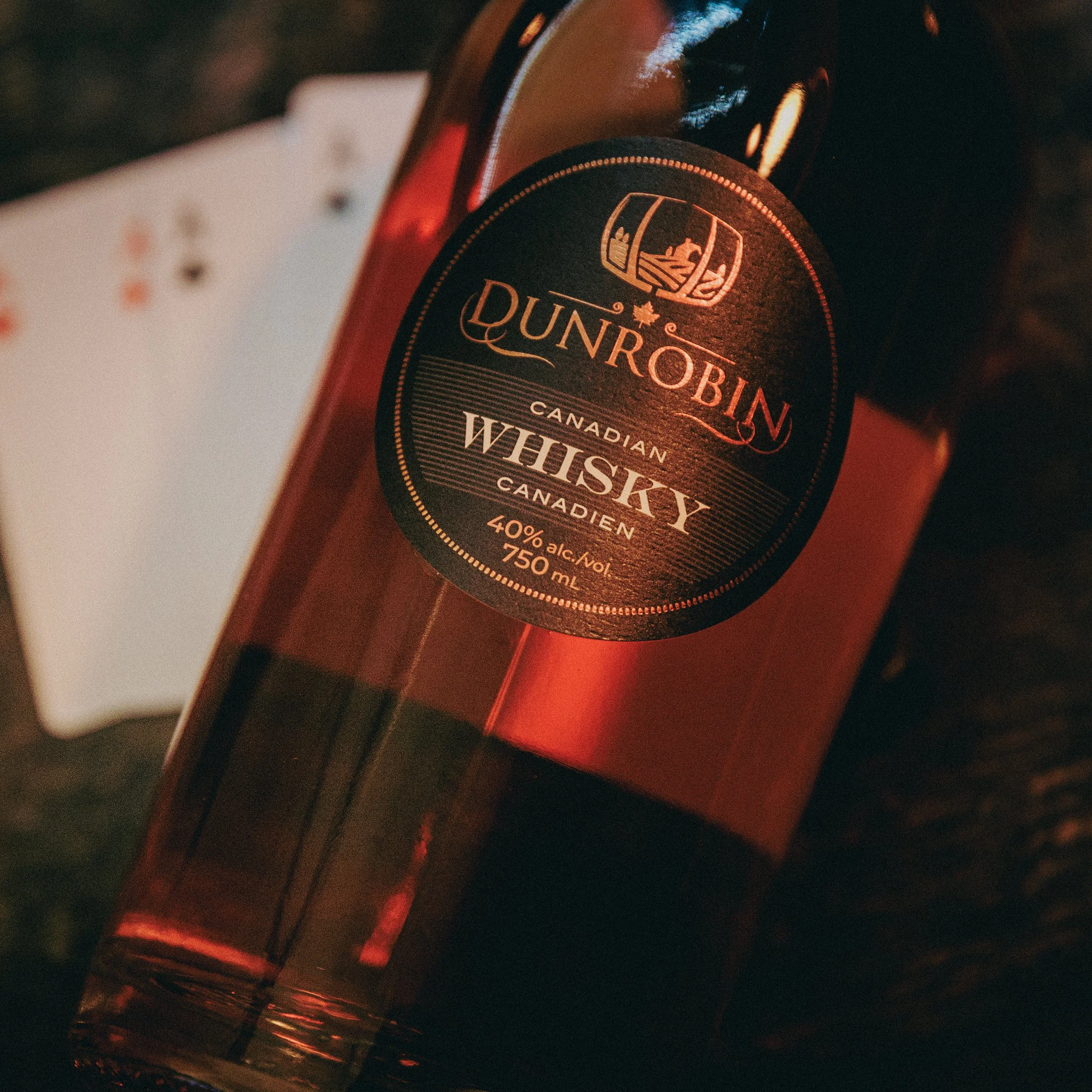 A bottle of Dunrobin Canadian whisky with a black and gold label, lying on its side on a dark surface. There is a playing card next to the bottle, partially visible.