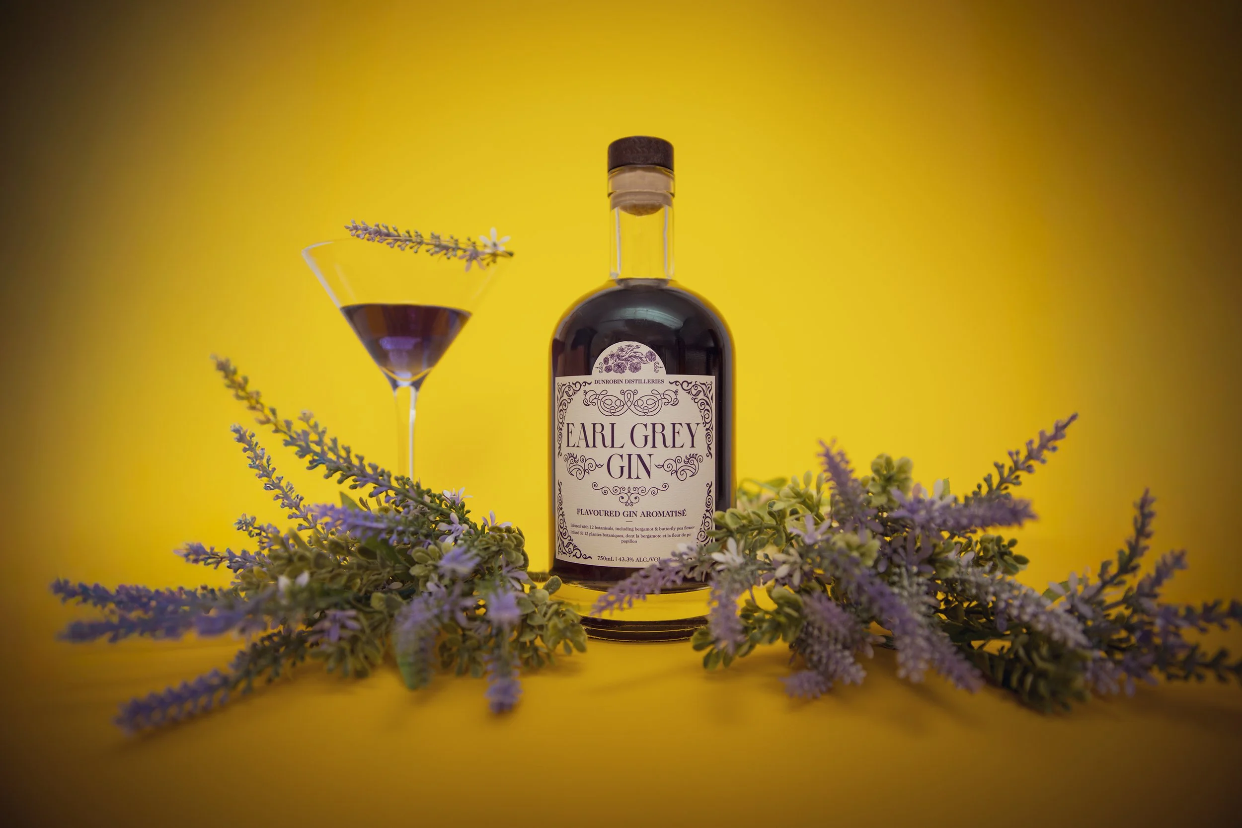 Bottle of Earl Grey Gin surrounded by purple lavender flowers against a yellow background, with a cocktail glass filled with purple liquid and lavender garnish on the left.