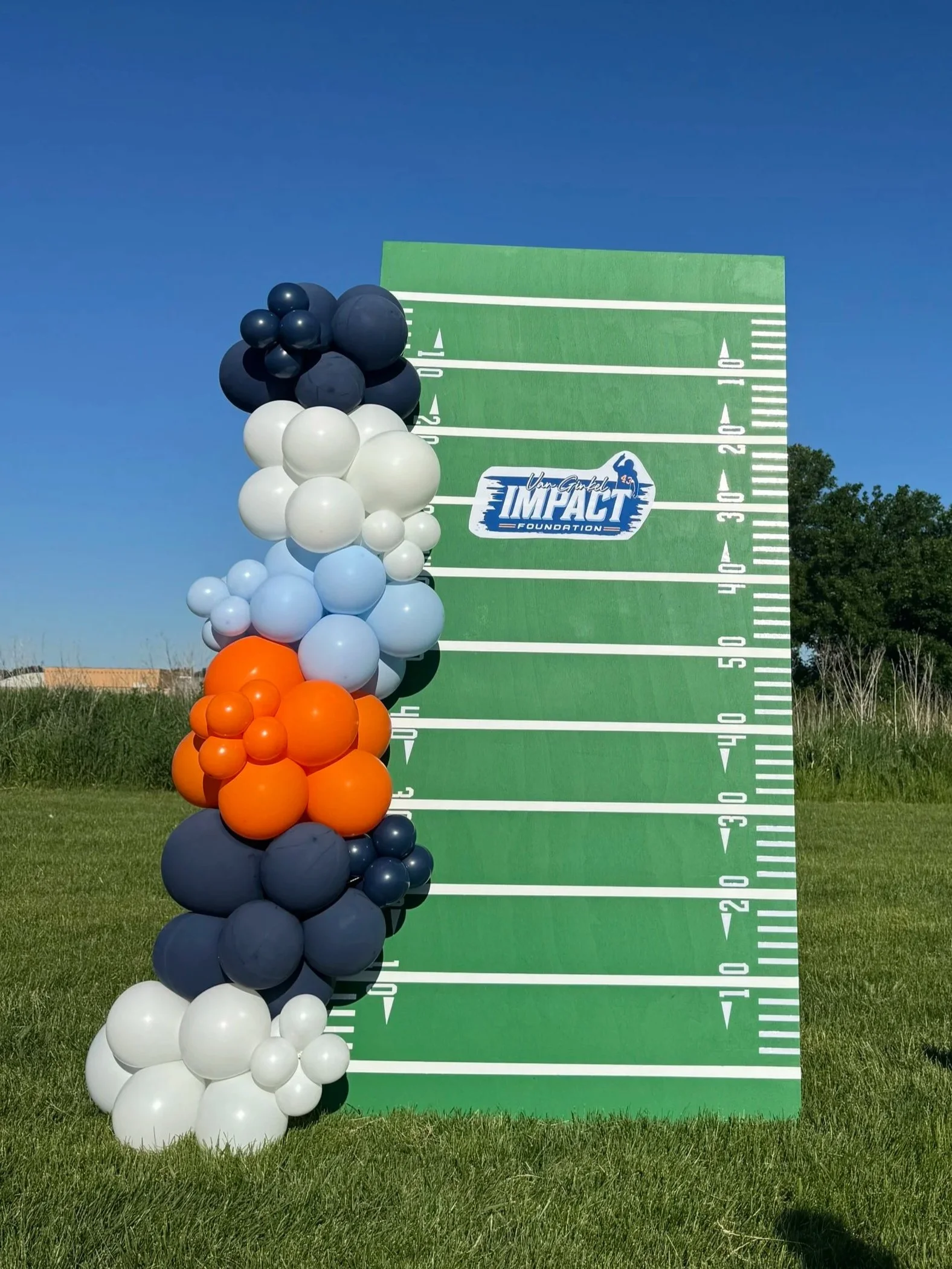 Large outdoor green football field display with the Impact Foundation logo, decorated with a cluster of black, white, orange, and gray balloons on a grassy field under a clear blue sky.