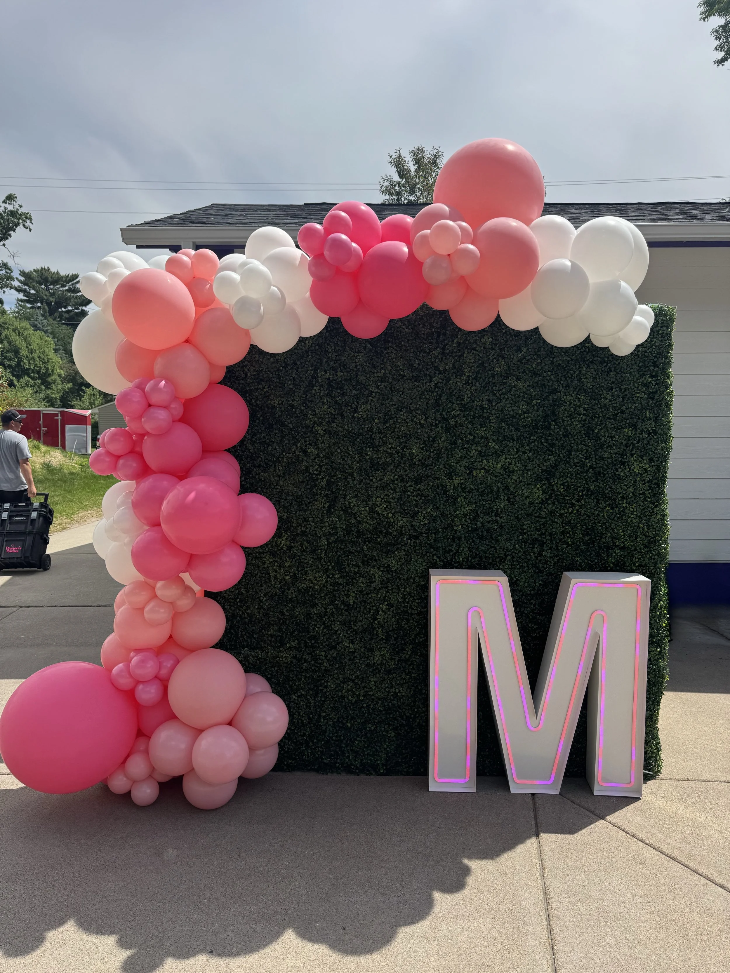 A decorated green hedge wall with gold, white, and green balloons arranged around it. A large illuminated number 25 is placed in front of the wall, indicating a celebration or birthday event.