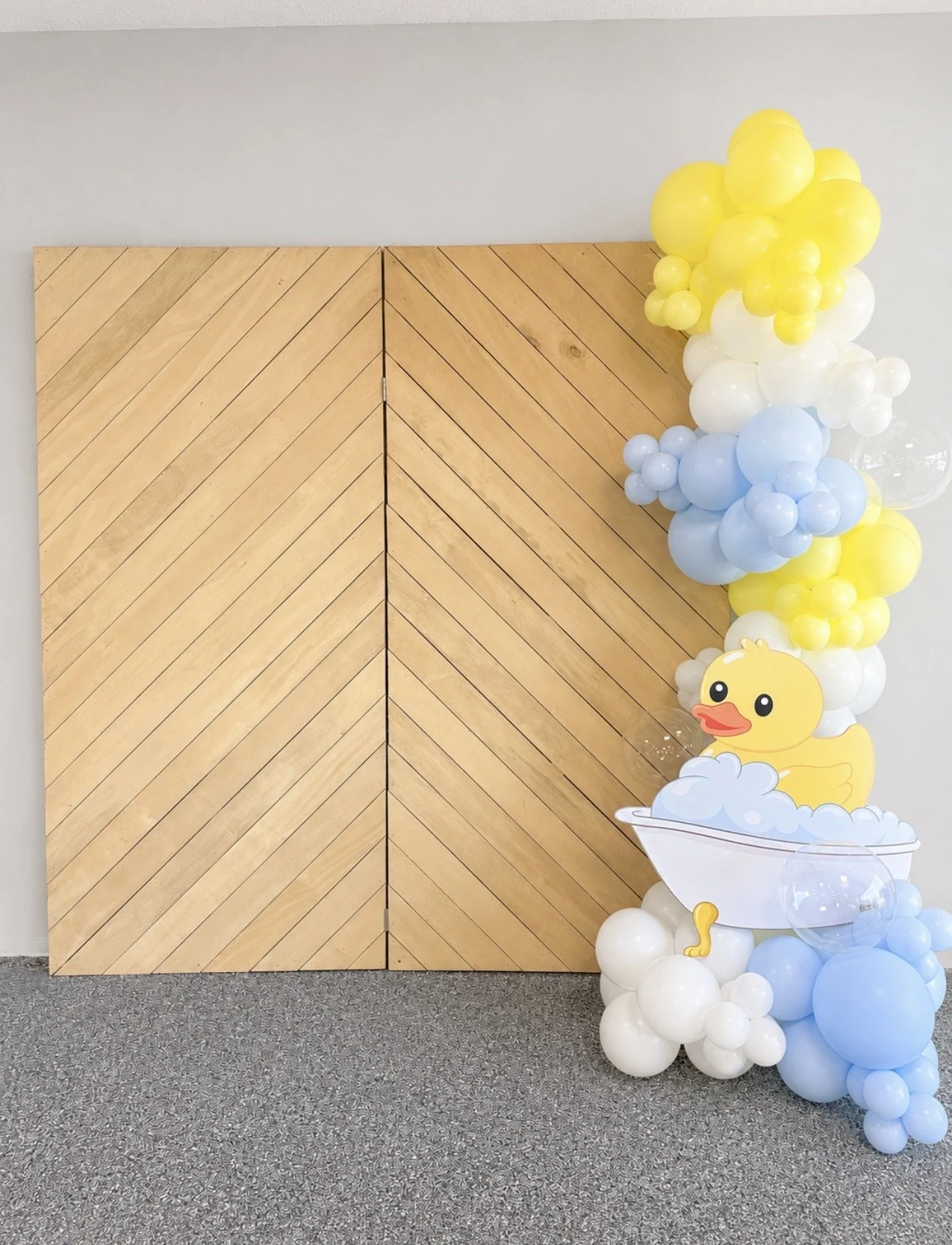 Decorative baby shower or birthday party backdrop featuring a wooden wall with a balloon banner in pastel yellow, white, and blue, including a cartoon rubber duck in a bathtub with bubbles.