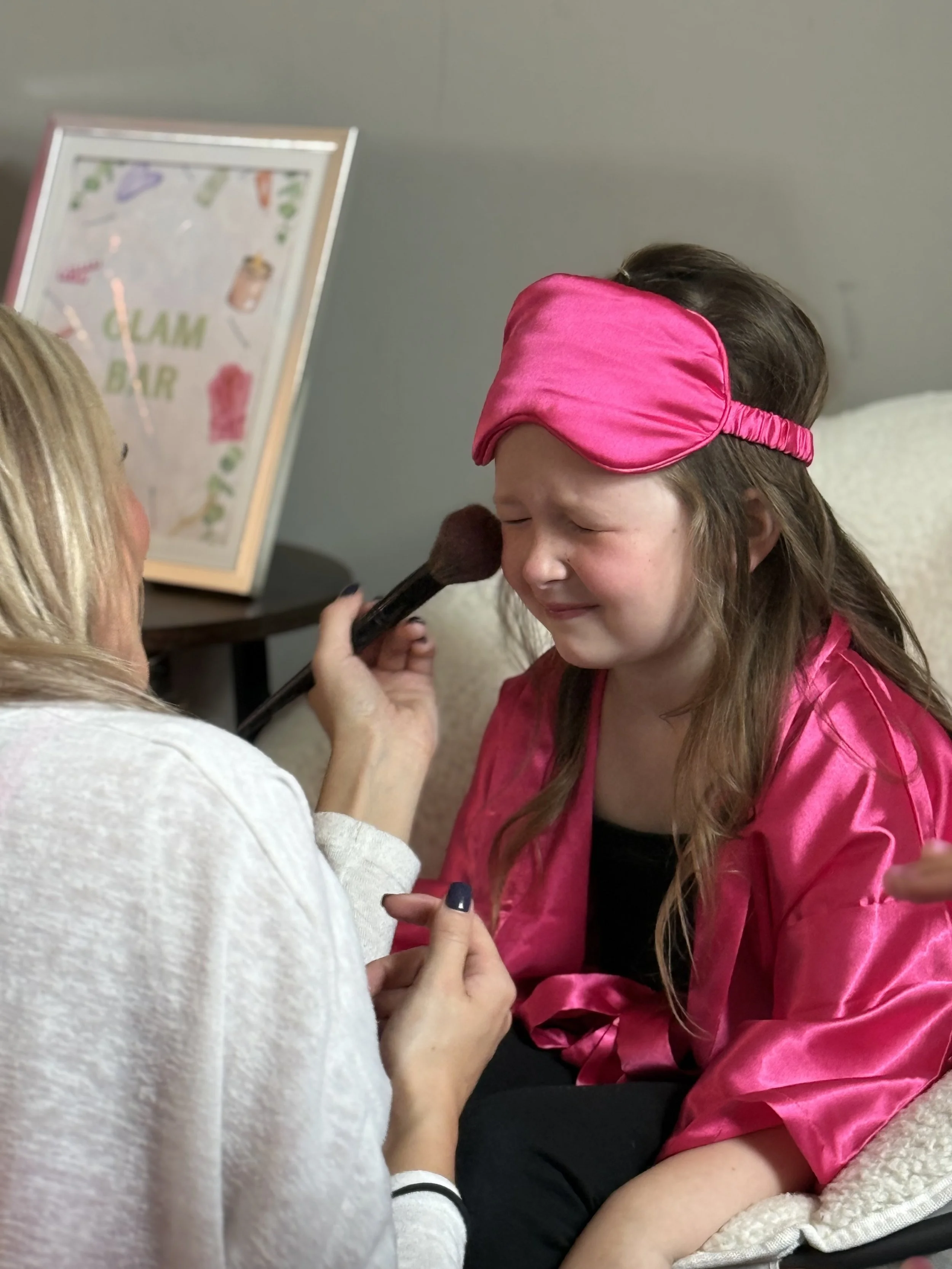 Child wearing a pink sleep mask, smiling, with a makeup brush near her face.