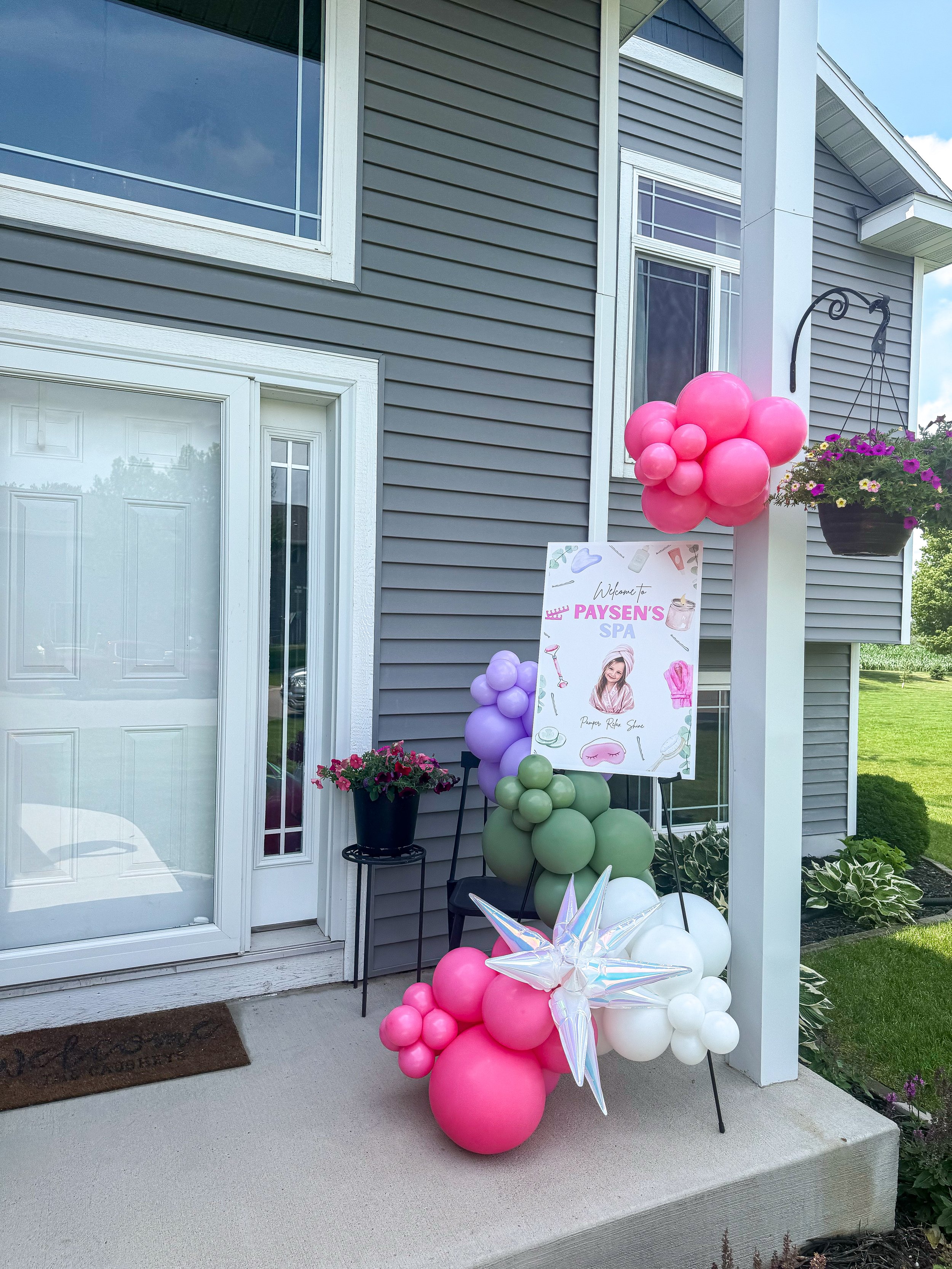Decorations for a baby shower or birthday celebration outside a house, including a large bunch of pink and white balloons, a sign with a photo of a young woman, and a hanging flower basket with purple and yellow flowers.