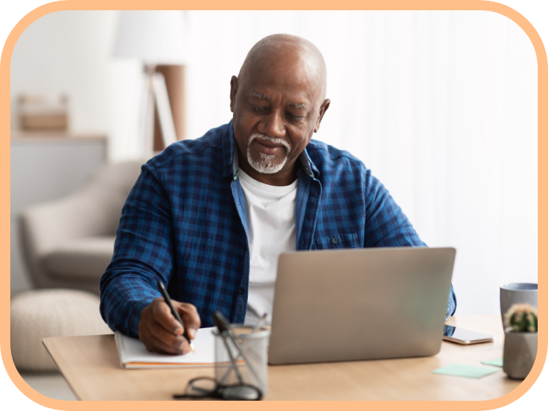 Older man writes in a notebook while using a laptop at a desk at home